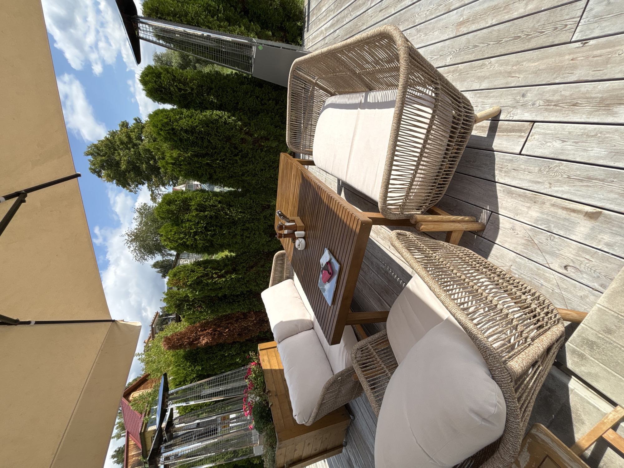 This image shows an outdoor seating area, possibly a patio or terrace, with a wooden table and several wicker chairs and sofas with light-colored cushions. On the table, there are salt and pepper shakers, cutlery, and a small tray with what appears to be a pink wallet and some cards. The seating area is set on a wooden deck. In the background, there are tall green trees, likely cypress or thuja, forming a dense hedge. To the left, a wooden planter box contains some pink flowers. Two tall patio heaters are visible, one on the left and another on the right, behind the hedge. A large beige umbrella provides shade over the seating area. A red-tiled roof of a building is visible in the upper left corner, peeking through the trees. The sky is blue with scattered white clouds. The scene suggests a restaurant or cafe setting in Zvenigorod, Russia, on a sunny day. There are no people visible in the image.