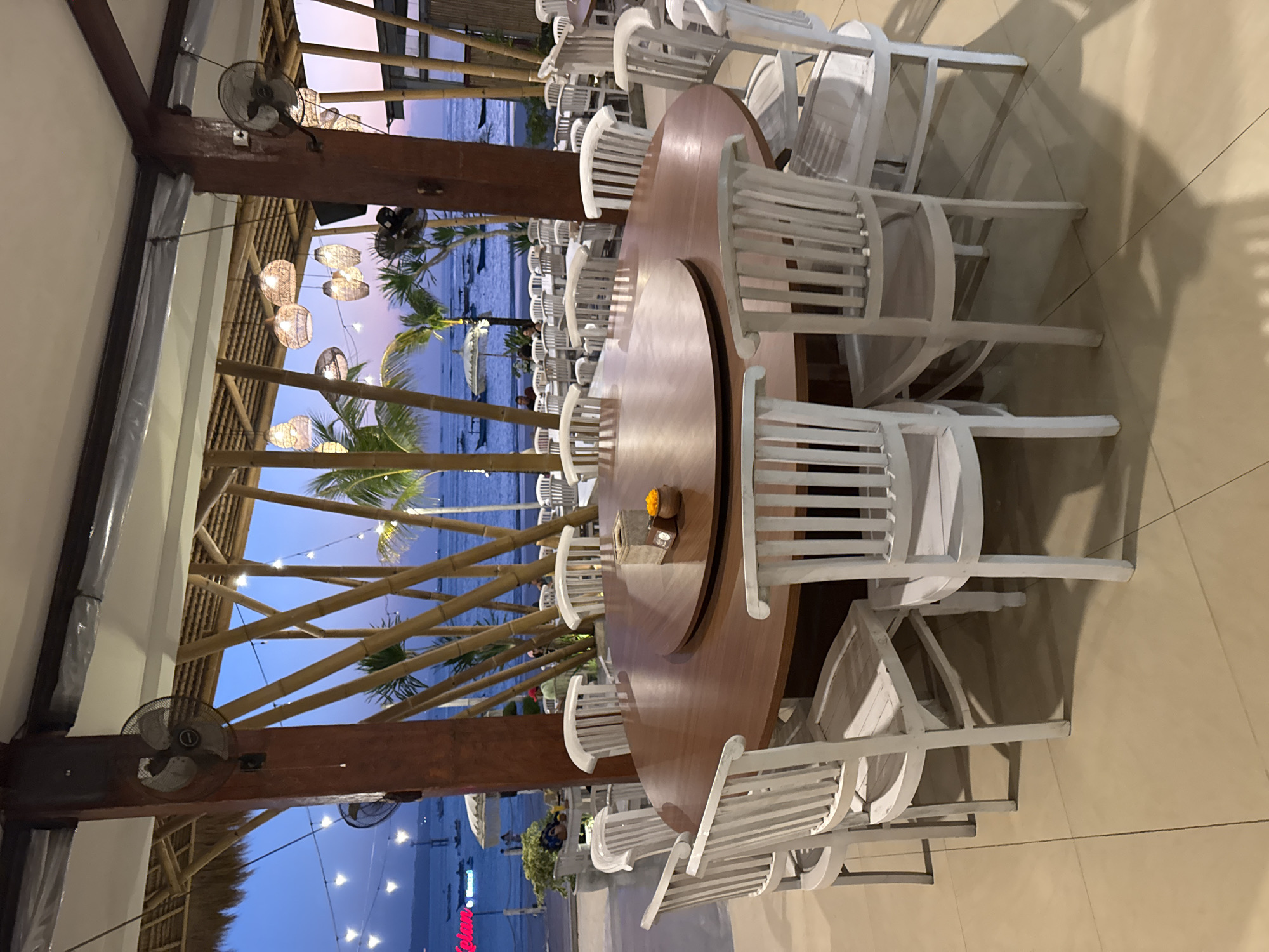 This image depicts a beachfront restaurant in Kuta, Indonesia, at dusk. A large, circular wooden table with a lazy susan sits in the foreground, surrounded by multiple white wooden chairs. The table is set with small decorative items. Beyond the table, the restaurant's open-air structure is visible, with a thatched roof and bamboo supports. In the background, the ocean stretches out to the horizon, with several small boats anchored in the water. The sky shows a gradient of blues and purples, indicating the transition from day to night. String lights are hung across the outdoor seating area, and a few palm trees are visible. The scene suggests a tranquil dining experience with a view of the sea.