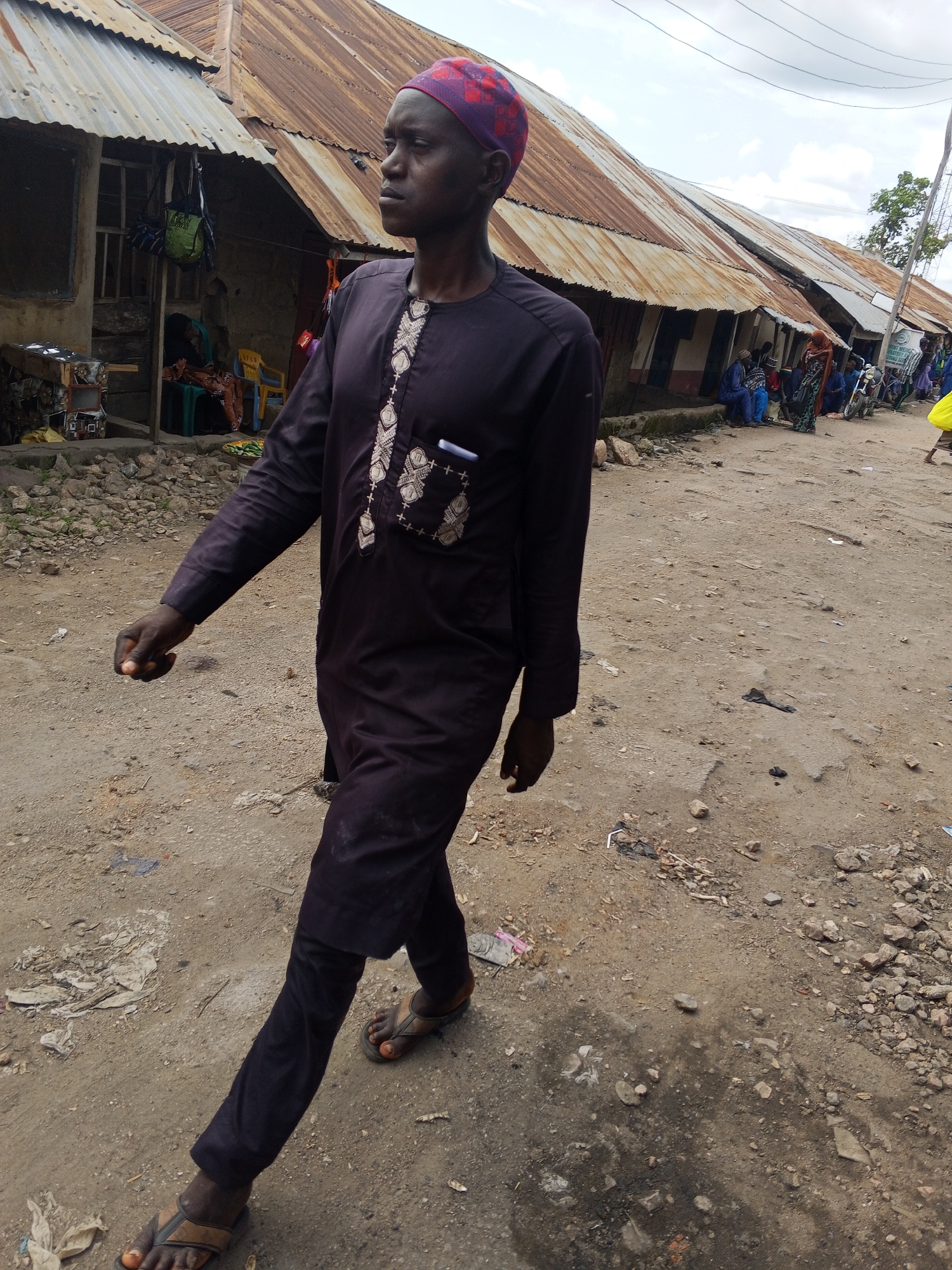 In Zango-ndaji, Nigeria, a young Black man wearing a purple kufi and a dark brown dashiki walks along a dirt road. The dashiki is adorned with white embroidery on the chest. He wears dark trousers and brown flip-flops. The man is captured mid-stride, looking to his left. Behind him, a row of structures with corrugated tin roofs lines the road. Several other people are visible in the background, some sitting and others standing near the buildings. A motorcycle is parked to the right. The ground is a mix of dirt, small stones, and scattered debris. The sky is overcast with clouds.