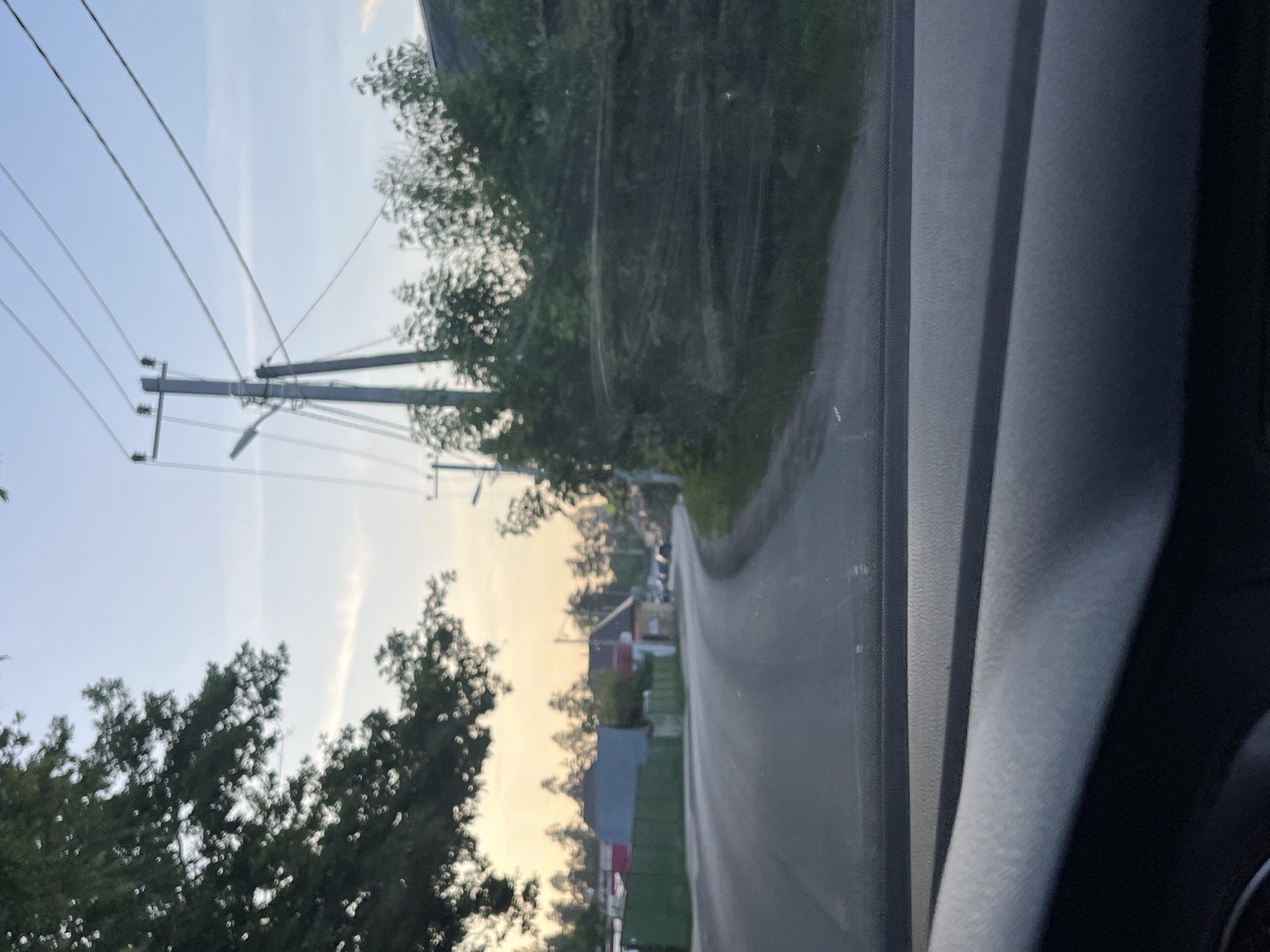 The image depicts a slightly blurred view from inside a vehicle, looking out onto a paved road that curves to the right. The road is bordered on the left by a green fence and some houses. On the right side of the road, there are trees and more houses, with power lines and utility poles visible against a pale blue sky with a hint of sunset orange on the horizon. The foreground of the image shows the dashboard of the car. The scene appears to be set in a rural or suburban area, possibly the city of Vasilyevo, Russia, given the architectural style of the houses and the presence of overhead power lines. There are no people clearly visible in the image. The overall impression is that of a drive through a residential area during the late afternoon or early evening.
