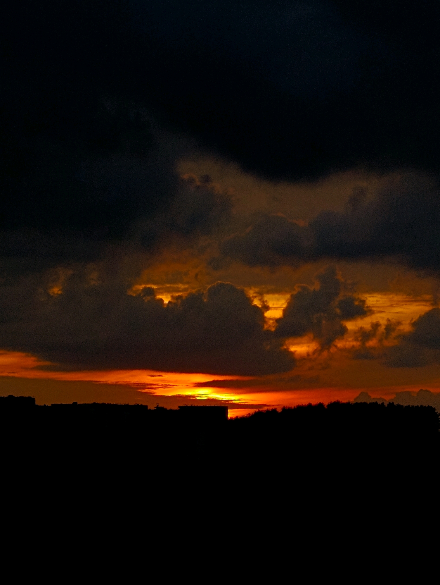 A silhouette of buildings and trees is visible against a dramatic sunset. The sky is filled with dark, heavy clouds that are illuminated from behind by a vibrant orange and yellow glow from the setting sun. The light is brightest at the horizon, with shades of red and deep orange radiating outwards. The buildings appear as dark, blocky shapes, and a line of trees can be seen on the right side of the image, also in silhouette. The overall scene is one of a city at dusk, with the natural beauty of the sunset contrasting with the urban landscape.