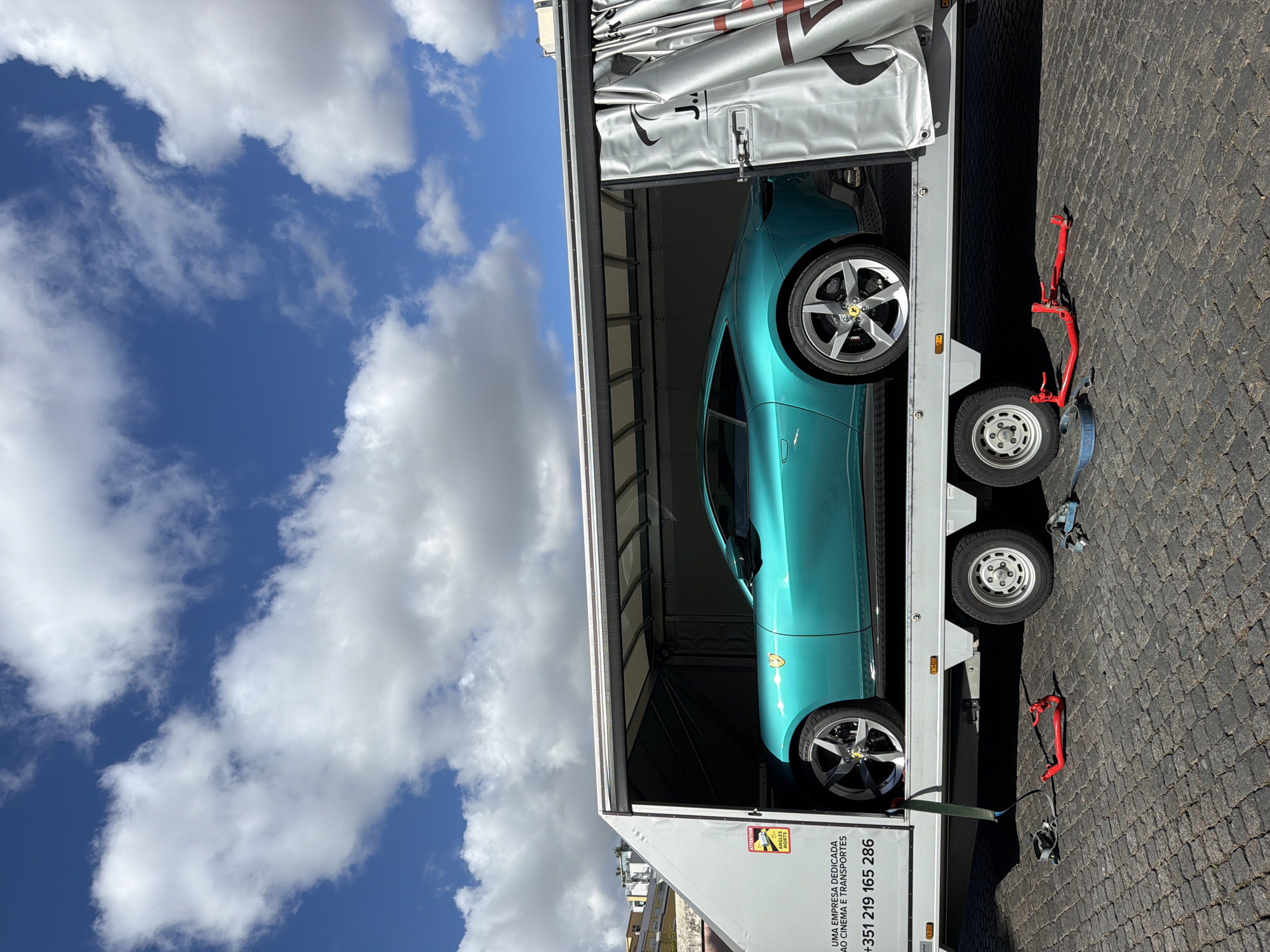 The image shows a light blue Ferrari inside an enclosed car carrier trailer. The trailer is parked on a cobblestone street. The sky is blue with scattered white clouds. The side of the trailer has text indicating a company dedicated to cinema and transportation with the phone number +351 219 165 286. Based on the phone number country code and street pattern, the location is likely Lisbon, Portugal.