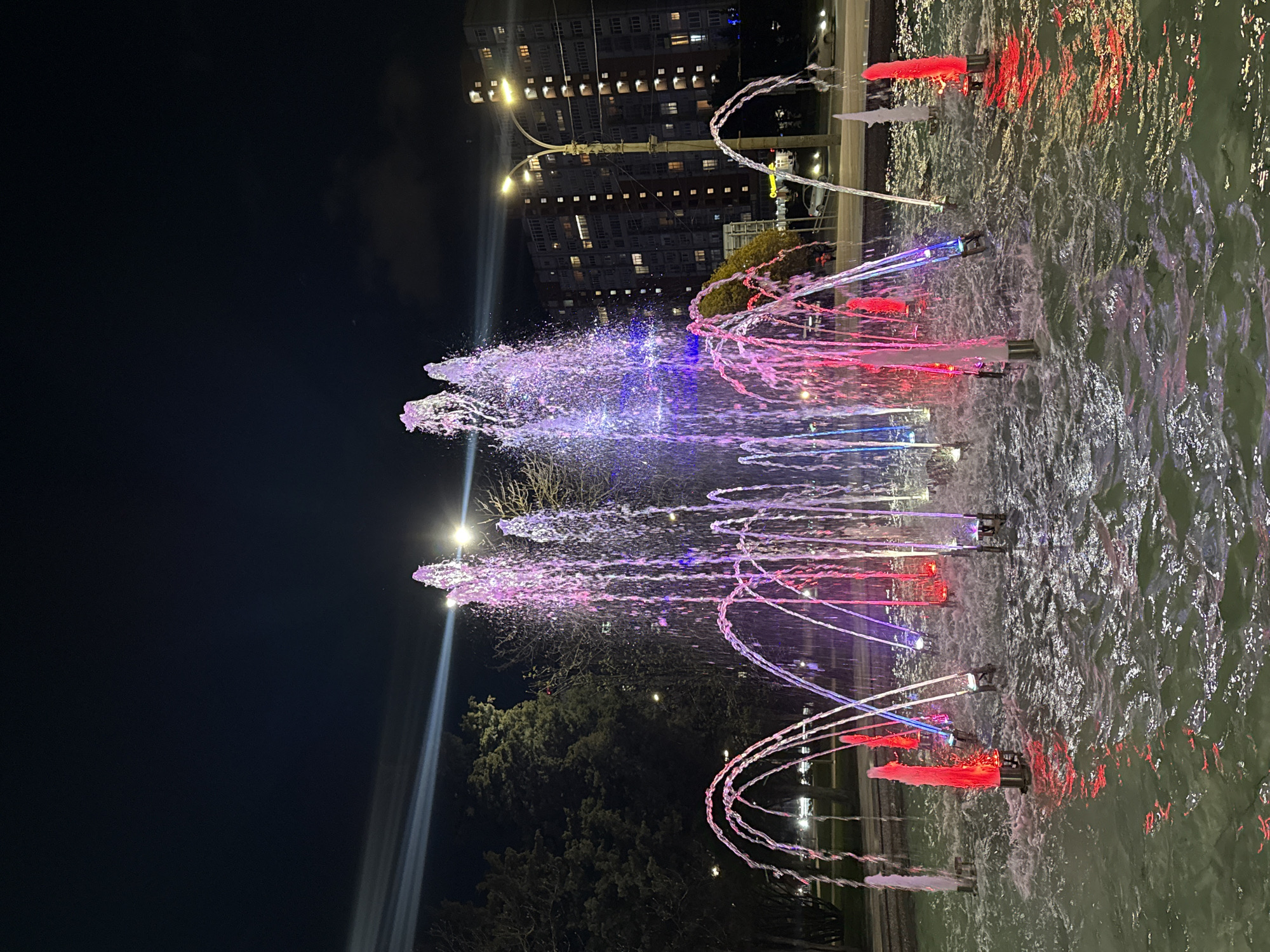 The image shows a fountain in Rostov-on-Don, Russia, at night. The fountain features multiple jets of water illuminated by colorful lights, including red, blue, and purple. In the background, there is a multi-story building with numerous lighted windows and a street light. Trees are also visible in the background. The water in the fountain has a greenish tinge.