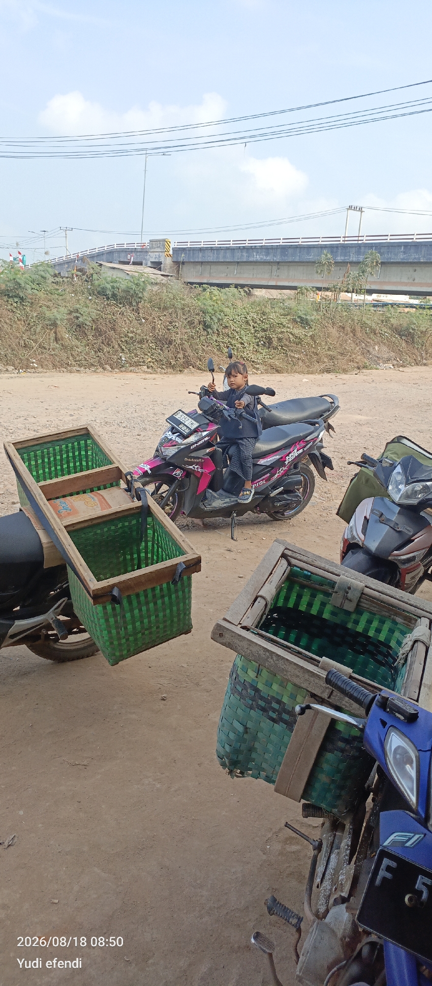 In Rawajitu Selatan, Indonesia, on August 18, 2026, at 08:50, a young girl sits on a pink and black motorcycle. The motorcycle is equipped with two green woven baskets attached to the rear, one on each side, and a wooden frame holding a box on the left side. Another motorcycle, mostly blue, is parked in the foreground on the right, also fitted with a green woven basket. The ground is dusty and dirt-covered, suggesting an outdoor, possibly rural or roadside setting. In the background, a concrete overpass stretches across the scene, with vegetation and power lines visible. The sky is partly cloudy.