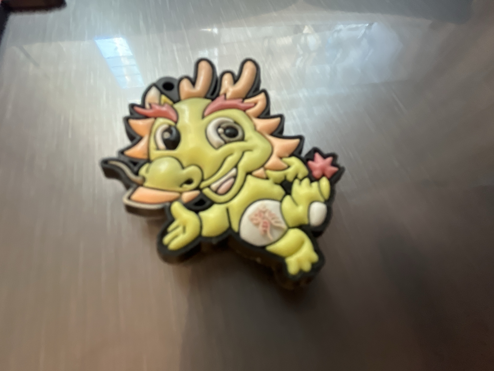 A close-up shot shows a cartoonish, yellow and orange dragon magnet adhered to a gray, possibly metallic surface. The dragon has black outlines and is depicted in a running pose with a happy expression. The surface reflects light, indicating a smooth, reflective material. The location and city are unknown.