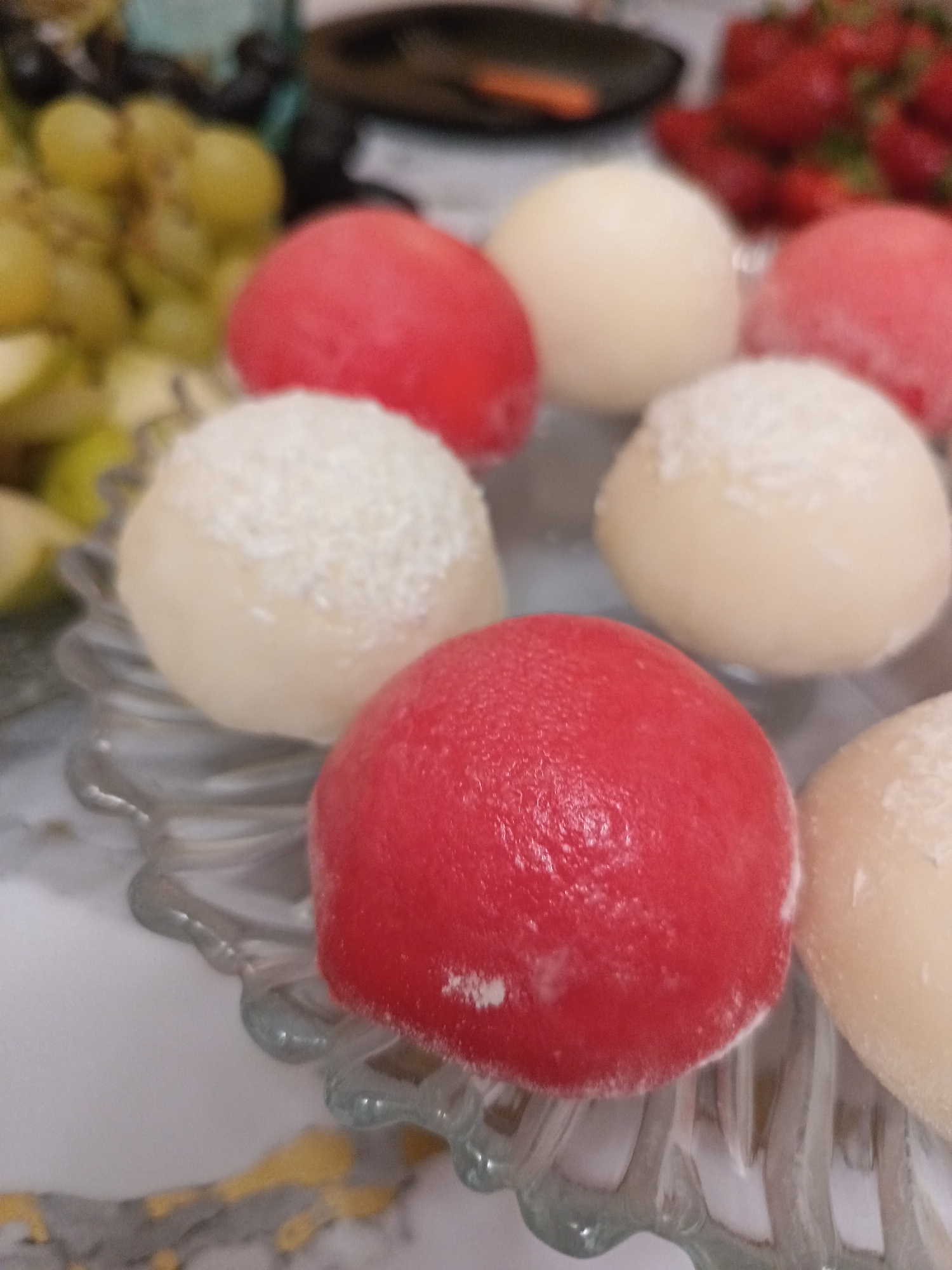 The image displays a close-up of several round, sweet pastries, possibly mochi or petit fours, arranged on a decorative glass plate. Some pastries are red, while others are a pale, creamy color, with some of the latter dusted with a white powder, resembling desiccated coconut. In the blurred background, there are clusters of green grapes, slices of apple, and a pile of red strawberries, suggesting a fruit platter or dessert spread. The overall setting appears to be a domestic environment, likely a kitchen or dining area. There is no indication of the city of Krasnodar, Russia, or any specific location within it. The scene is static and does not involve any people, actions, or interactions.
