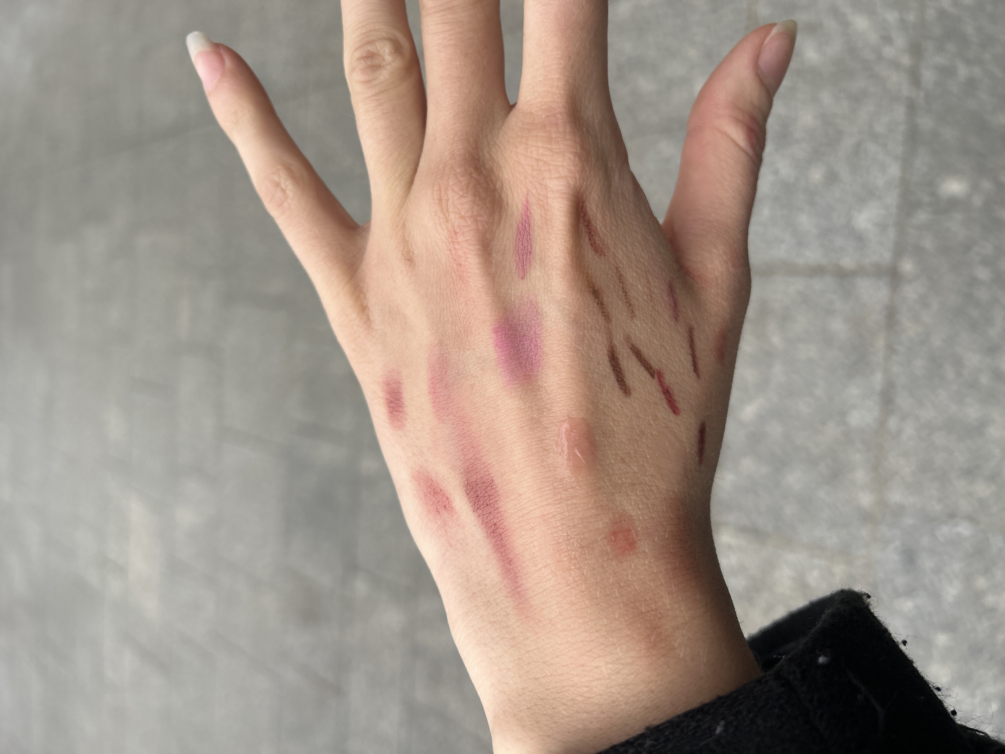 The image shows a fair-skinned human hand with various cosmetic product swatches on the back of the hand and fingers. The swatches include pink and brown shades, possibly lipsticks or lip liners. There is also a small, clear droplet on the back of the hand. The fingernails are long and unpainted. The lower right corner of the image shows a portion of a black garment. The background is a blurred gray, possibly a concrete or stone surface. The location in the city of Paris, France cannot be determined from the content.