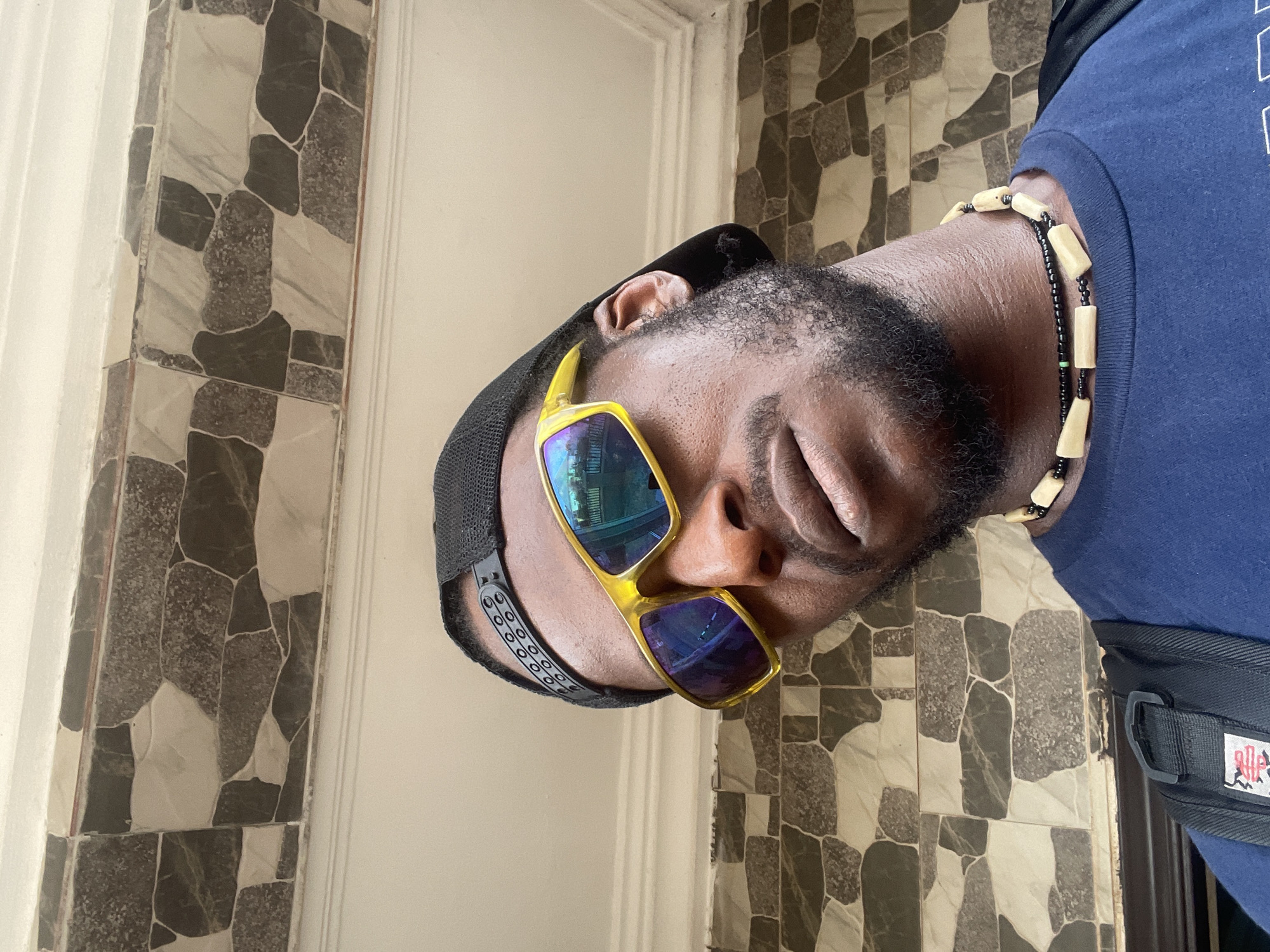 A close-up, sideways portrait shot features a dark-skinned individual with a short black beard and mustache, wearing a black cap or durag. The person's head is tilted significantly to their right (viewer's left). They are wearing distinctive yellow-framed, reflective sunglasses that obscure their eyes, showing blueish reflections of what appears to be an indoor or semi-enclosed environment. The person's expression, with the mouth slightly closed, appears neutral or contemplative.
A multi-stranded beaded bracelet or necklace adorns their arm or neck, featuring light wooden, rectangular beads interspersed with smaller black beads, and a small green bead is visible on one strand. They are dressed in a dark blue t-shirt, and a black strap of a bag or backpack with a white and red logo (illegible) is visible over their right shoulder. The background consists of a textured wall covered with irregularly shaped tiles in shades of grey, black, and off-white/cream. White door frames are visible on both the left and right edges of the composition, suggesting an indoor setting. The lighting indicates it is daytime. The image captures a personal moment, likely posed, in what appears to be an indoor location in Abuja, Nigeria.