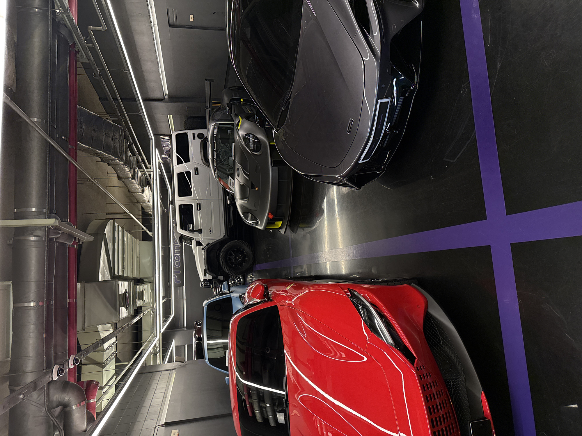 The image captures a display of luxury vehicles in a parking garage in Seoul, South Korea. The garage has a black floor with purple lane markers. The cars include a red Ferrari, a gray Porsche, a white Mercedes-Benz G-Wagon, and a dark gray McLaren. The garage has a ventilation system running along the ceiling.