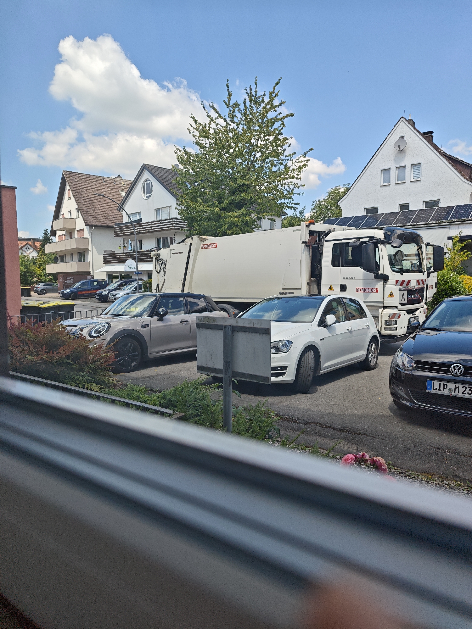 In Bad Meinberg, Germany, a refuse collection vehicle is parked on a street adjacent to residential buildings. The truck is white with a large, beige refuse body. A white passenger car and a silver hatchback are parked in front of the refuse truck. The buildings in the background feature red-tiled roofs and balconies. The sky is blue with scattered white clouds. A tree with green foliage is visible between the buildings.