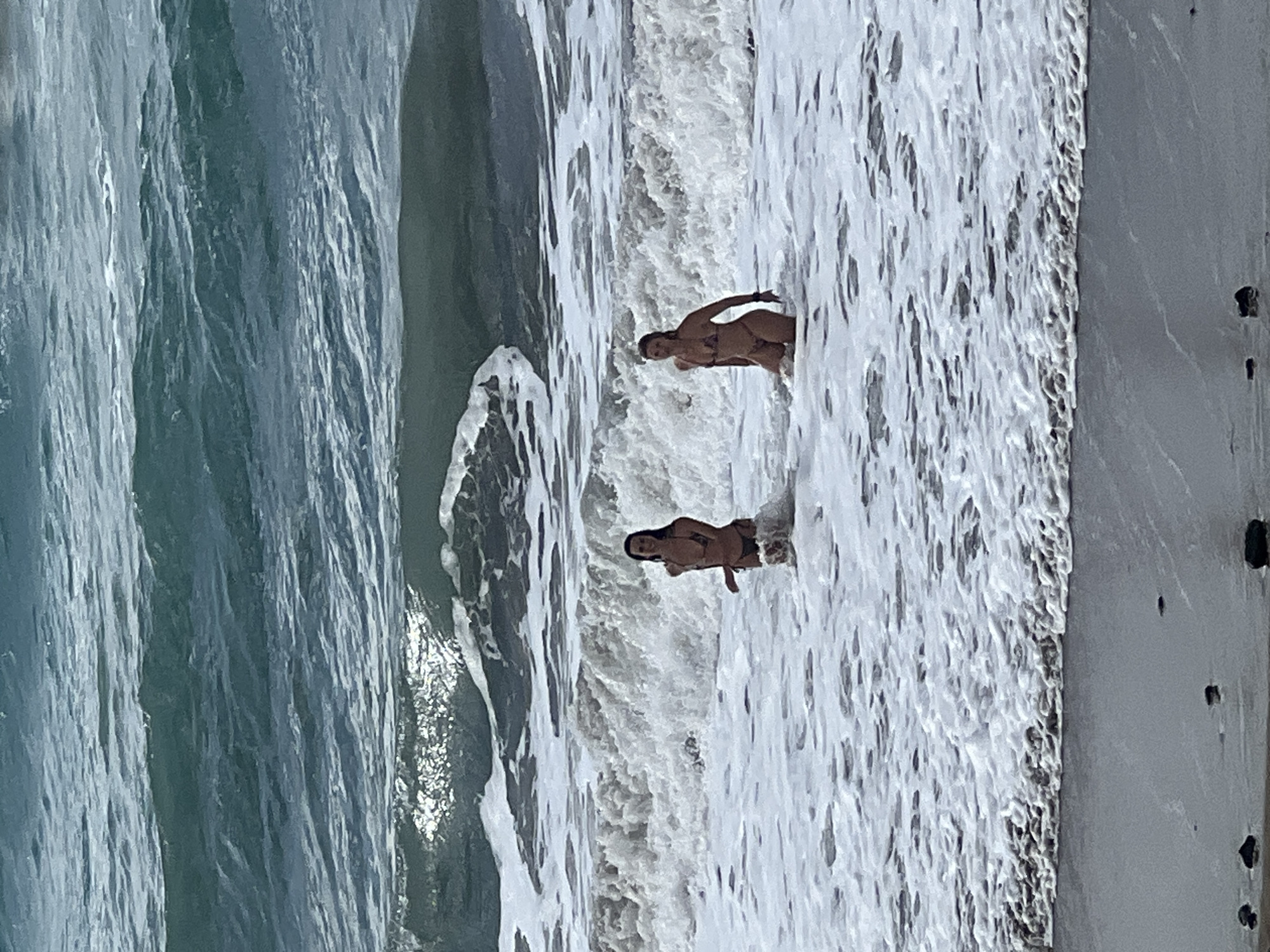 Two individuals, appearing to be women, are captured standing in the foamy white surf of an ocean wave. The image shows a beach scene on what appears to be a bright, sunny day in Las Palmas, Spain.
On the left side of the frame, the deeper blue and green tones of the open ocean are visible, indicating the body of water from which the waves are approaching. The middle section is dominated by the dynamic, turbulent white foam of breaking waves, where the two subjects are positioned. They are both wearing two-piece swimsuits and are partially submerged in the water, with the foam reaching roughly to their waist or thighs. One person is slightly further into the water than the other, who stands closer to the shore. To the right, a darker, wet stretch of sand marks the immediate shoreline. The overall atmosphere suggests a leisurely activity, enjoying the ocean and beach environment. No text is visible in the image.