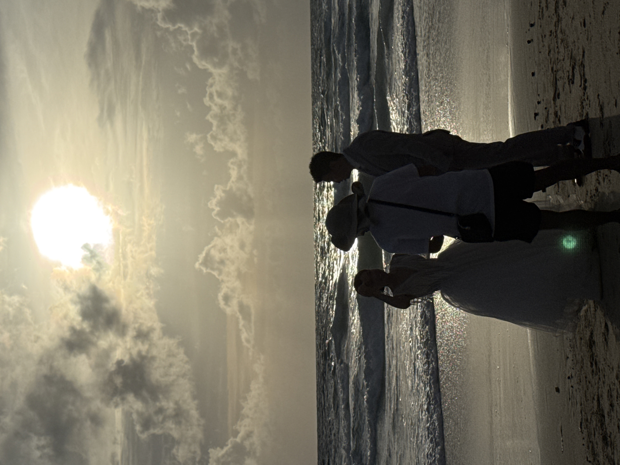 On a beach in Karon, Thailand, a wedding photoshoot is in progress. The sun is high in the sky, partially obscured by clouds, casting a golden light over the scene. A bride in a white gown stands on the left, facing away from the camera, holding her hand up to her head. To her right, a groom in a light-colored suit stands facing her, looking towards the ocean. Between the couple and the camera stands a photographer, wearing a hat and a dark bag slung across their body, with their back to the camera. The ocean is calm with gentle waves lapping at the shore. The sand is dark and wet near the water's edge, with a few scattered pebbles. The sky is a mix of light clouds and clear patches, reflecting the sun's glow.