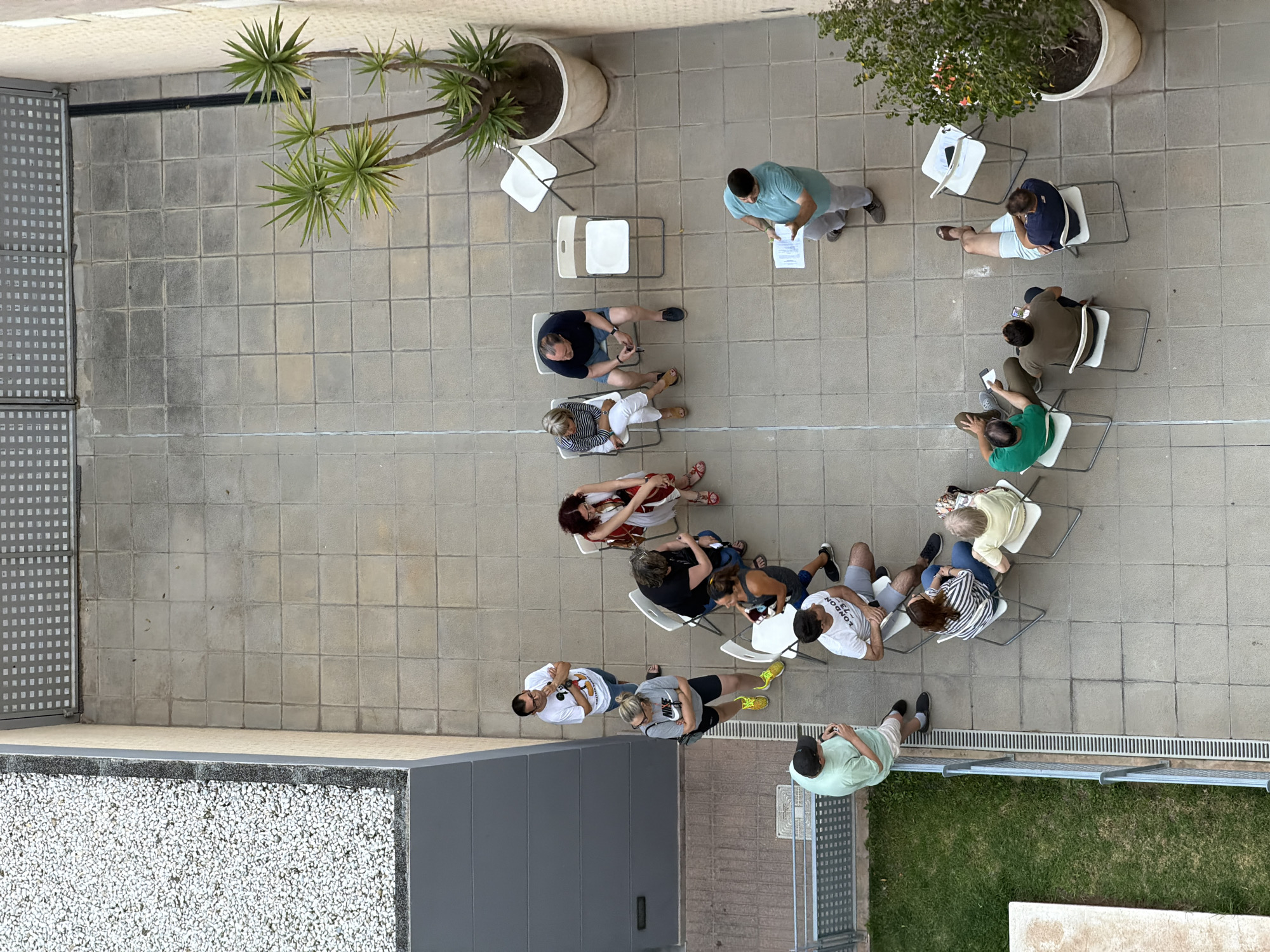 The image depicts an outdoor gathering of approximately 15 people seated and standing on a paved courtyard in Benimàmet-beniferri, Spain. The courtyard is tiled with square, light-colored pavers. To the left of the courtyard, there is a building with a gray, textured wall and a section of white gravel. A metal railing and a grassy area are visible along the edge of the courtyard. In the background, a building with a gray, perforated facade is present. Several white folding chairs are arranged in a loose circle, with some people seated in them and others standing. One person, dressed in a teal polo shirt and light gray pants, is standing and holding a document, seemingly addressing the group. Another individual, wearing a light blue t-shirt and shorts, is also standing and looking towards the speaker. The remaining individuals are seated in chairs, observing the speaker or interacting amongst themselves. A potted plant with spiky green leaves is visible on the right side of the courtyard, and another smaller potted plant with colorful flowers is near the bottom right. The scene appears to be a casual meeting or presentation in a residential or community outdoor space.