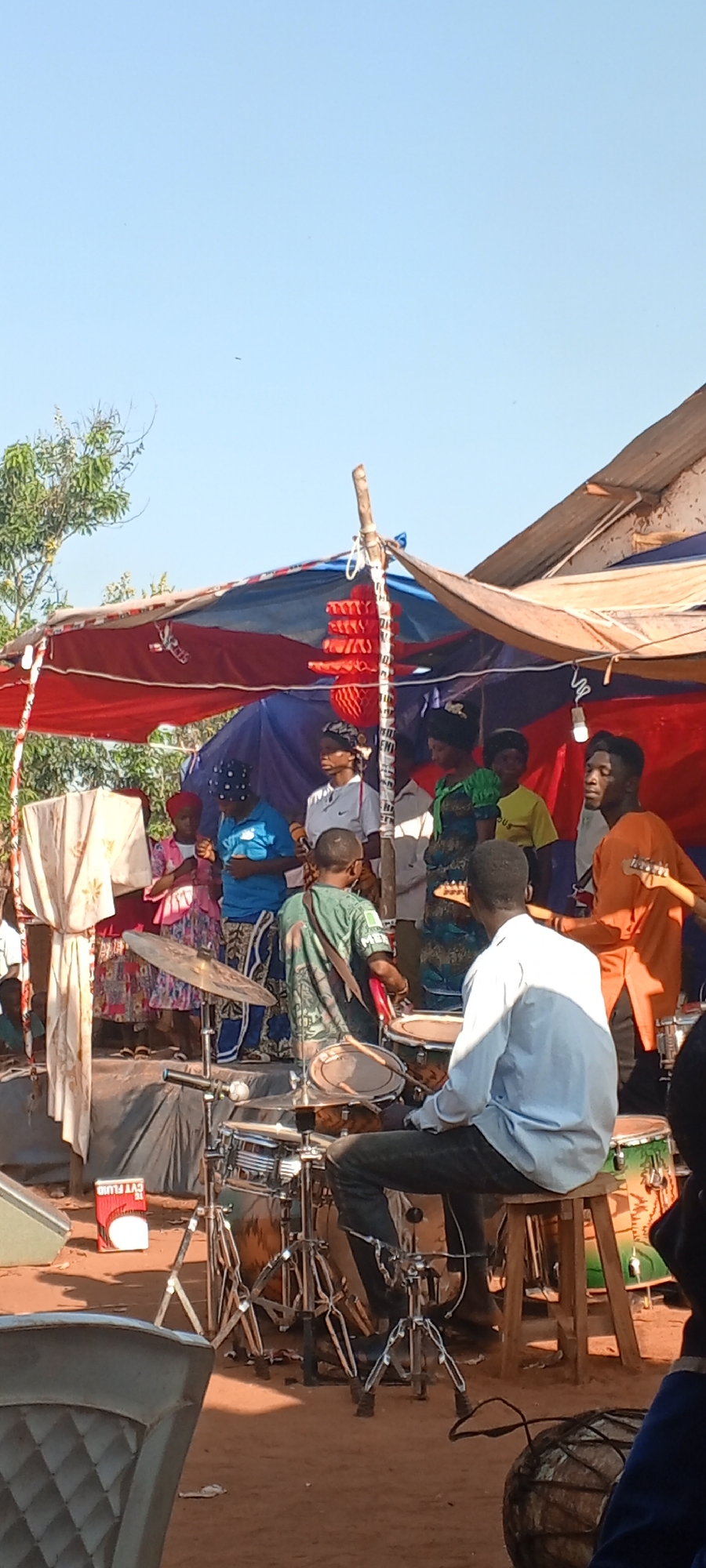 A band is performing on a makeshift stage in Ngalula, Democratic Republic of the Congo. The stage is covered by a red, white, and blue tarp supported by wooden poles. A group of people are gathered on the stage, some of whom appear to be singing. A drummer sits in front of his kit, while a guitarist stands to the right of the stage. A can of "CVT FLUID" is visible near the front of the stage on the left, and a wooden stool supports the drummer. The ground is dirt.