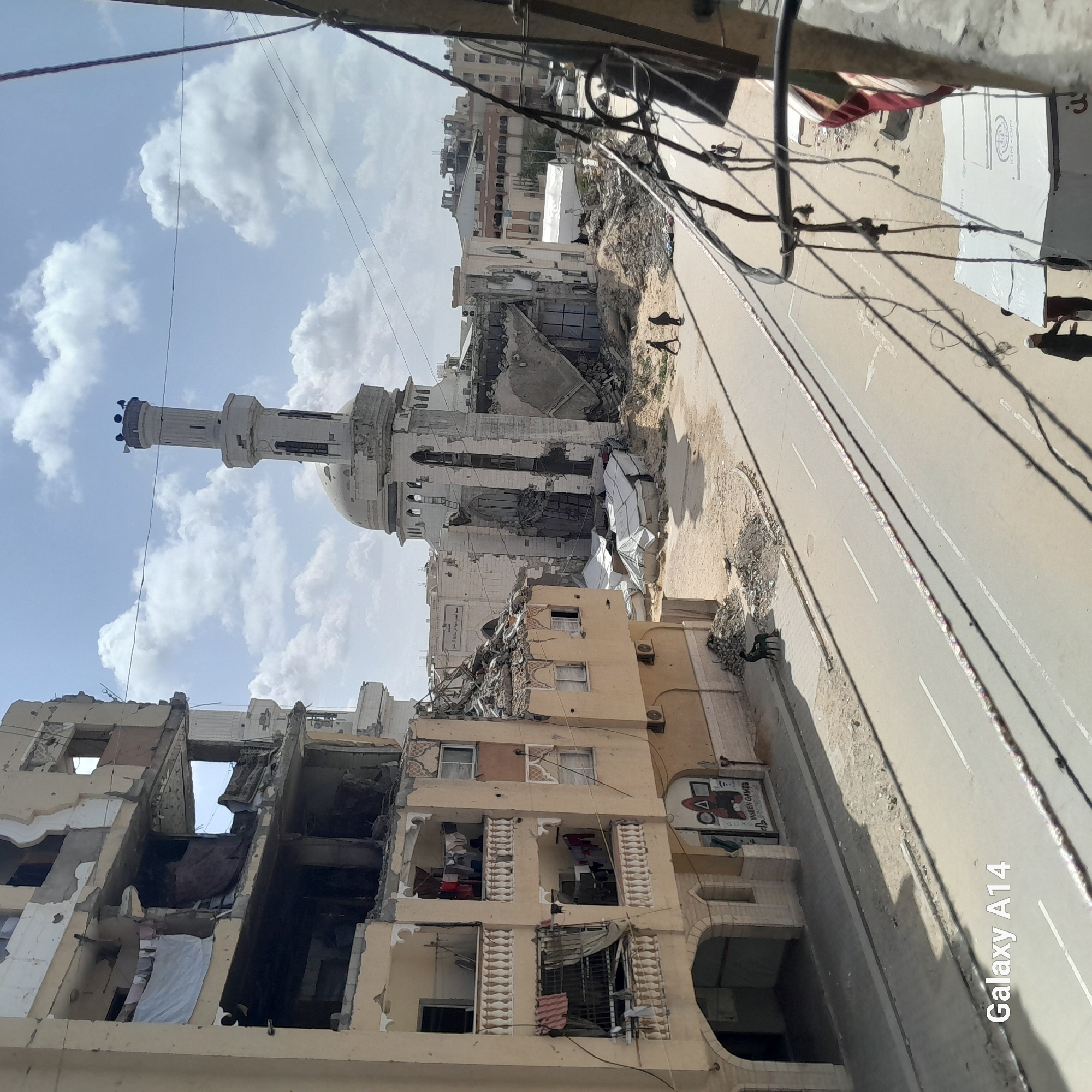 The image shows significant destruction in a city. Buildings are heavily damaged, some with gaping holes and collapsed structures. A mosque with a tall minaret stands amidst the ruins. The street is lined with rubble. There are also tents set up in the area. Two people walk along the street. Overhead, power lines are visible.