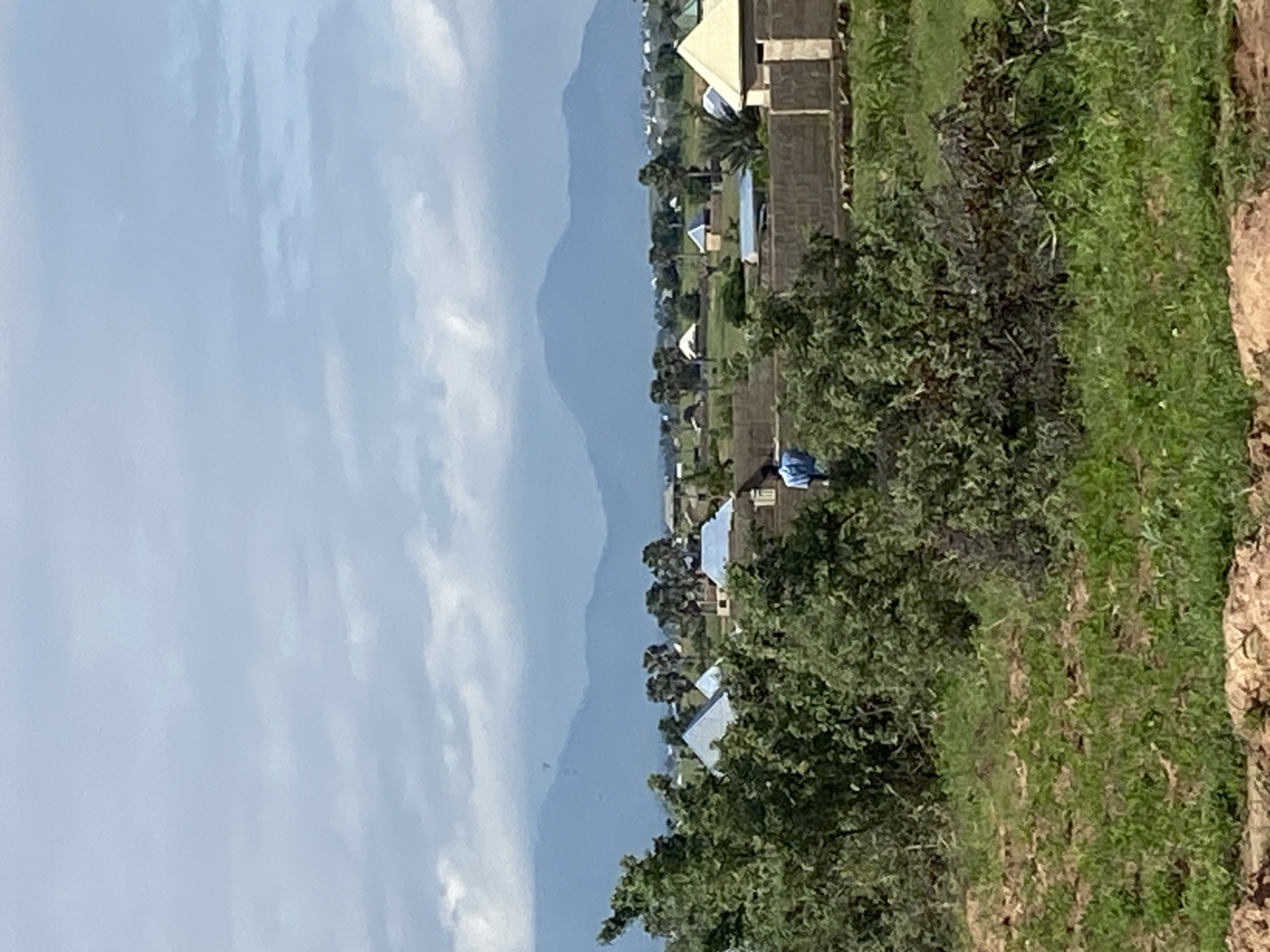 The image shows a wide shot of Kusan, Nigeria, under a cloudy sky. In the foreground, there is green grass and some rocky terrain. Beyond that, there are lush green trees and bushes. Behind the trees, there is a village with houses that have conical or pyramidal roofs. Some of the houses are constructed with brick walls. In the background, there are mountains in the distance. There is one person visible in the image, wearing a blue shirt and standing near a brick wall. The overall scene is of a rural landscape in Nigeria.