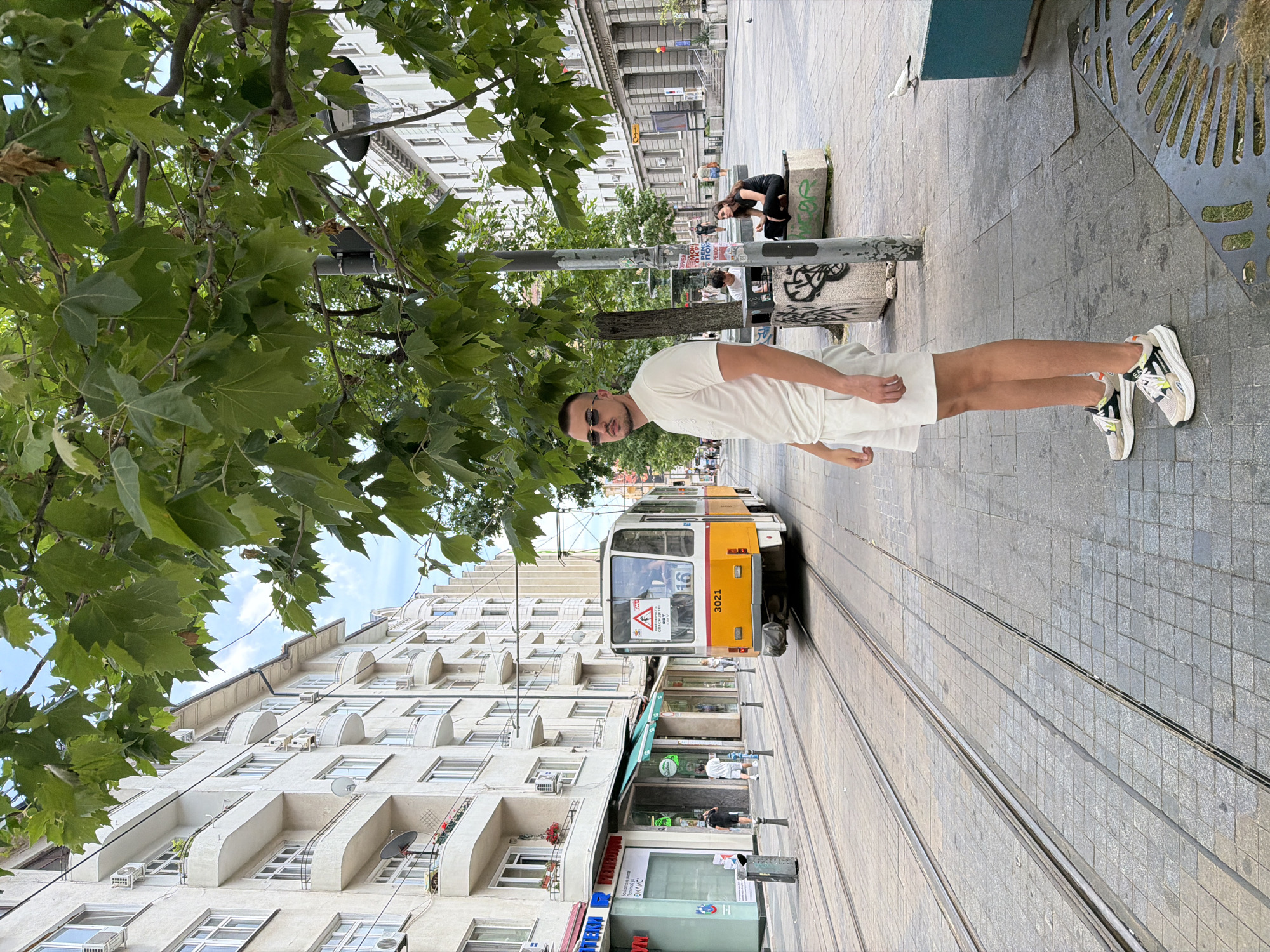 A man is standing on a sidewalk in front of a tram. The tram is yellow and white, with the number 3021 visible on the side. The man is wearing a white t-shirt and white shorts, and he has on sunglasses. He is looking to his left. Behind him, there is a tram track and a street. To the left of the tram track, there are buildings with shops. To the right of the tram track, there is a street with more buildings and trees. Two people are sitting on a bench in the background, to the right of the man. The scene appears to be on Vitosha Boulevard in Sofia, Bulgaria.