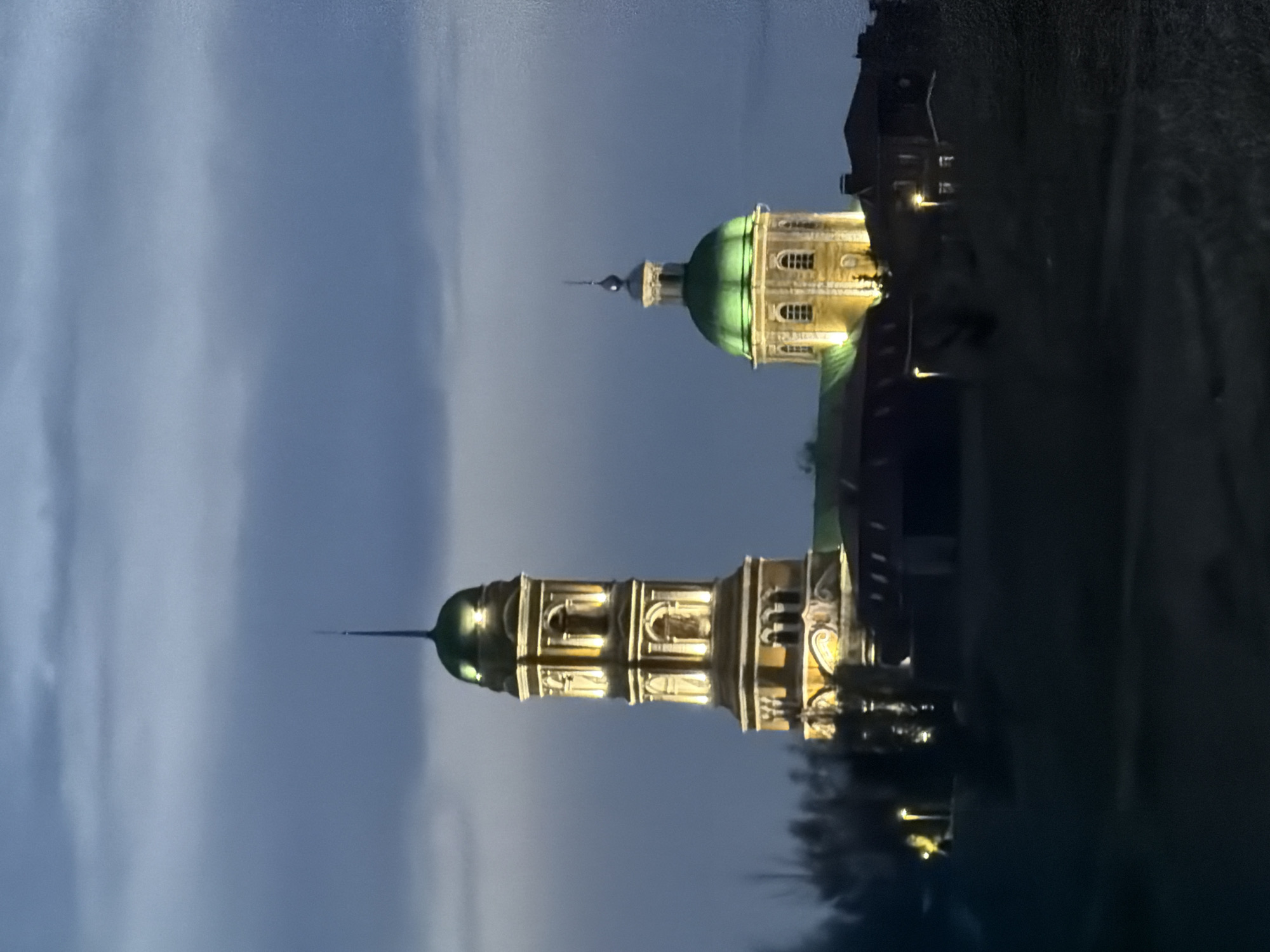 The image shows the illuminated domes and upper portions of the Church of St. Nicholas in Nikolo-Beryozovka, Russia, at dusk or night. The sky is a gradient of blues and grays, suggesting twilight. The church structures are lit up, showcasing their architecture. Dark vegetation or trees are visible on the right, contrasting with the lighter sky and illuminated buildings.