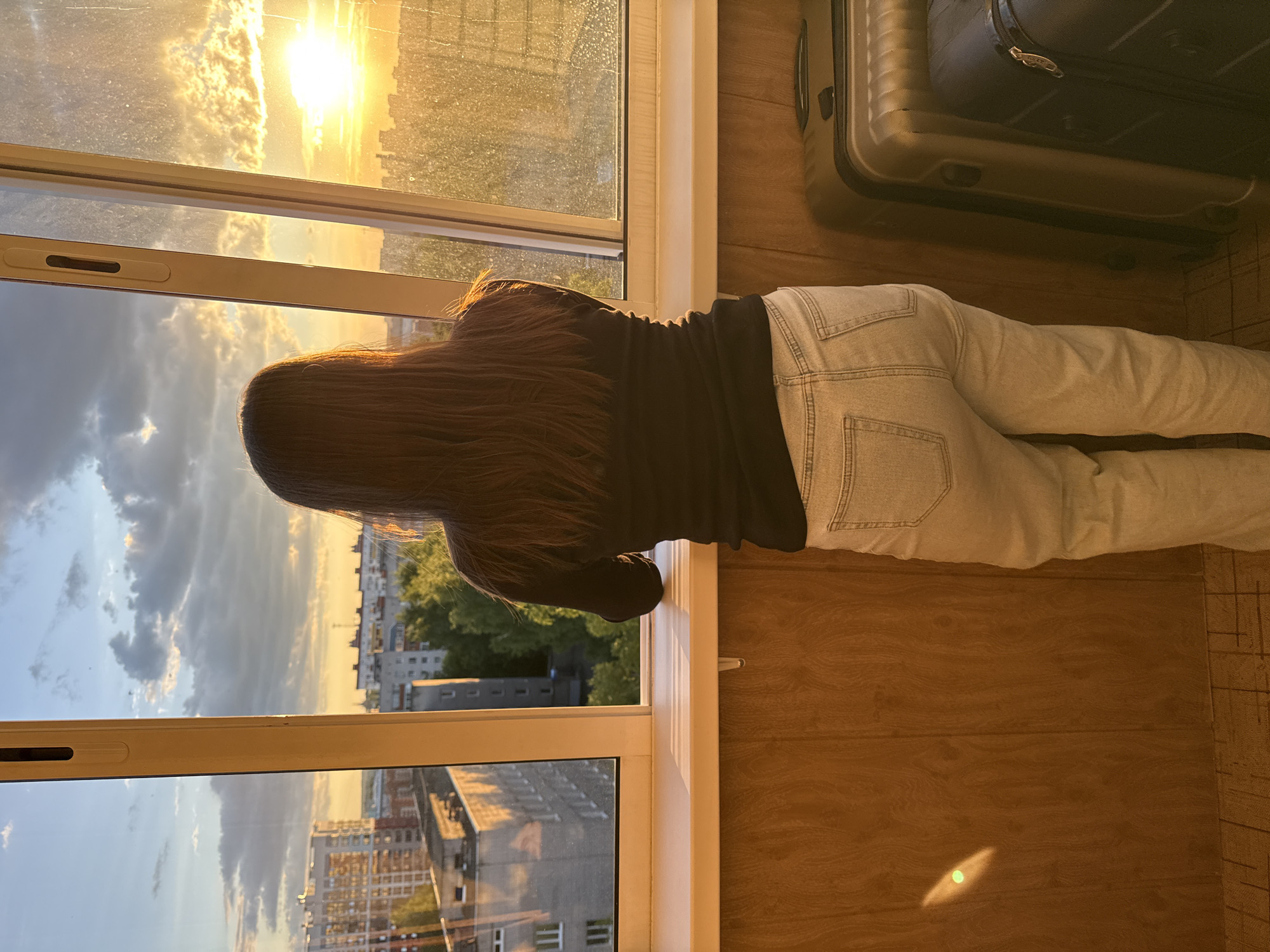 A person is depicted lying on their stomach, gazing out of a large window towards an urban landscape, likely in Izhevsk, Russia. The individual has long, dark brown hair and is dressed in a black long-sleeved top and light-colored denim jeans. Their head is positioned near the white window sill, looking outwards.
Outside the window, an urban environment with numerous buildings and some distant trees is visible. The sky is marked by partial cloud cover, but the prominent feature is the bright, low sun, which is setting or rising, casting a vibrant golden-orange light across the clouds and the horizon. This warm light illuminates the window frame and highlights parts of the person's hair.
Inside the room, the floor is covered with light brown wooden planks. Two suitcases are visible near the person's feet: one dark grey/black and another lighter, possibly metallic tan. The scene suggests a moment of relaxation or contemplation, enjoying the view during the golden hour. No visible text is present in the image.