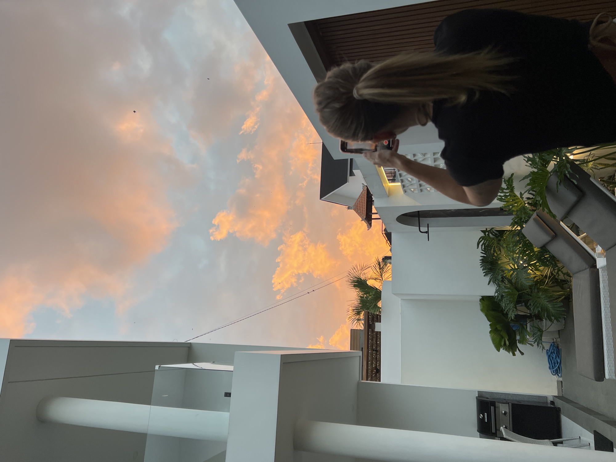 A woman is capturing a sunset with her phone. The sky is painted with vibrant orange and pink clouds, creating a dramatic backdrop. The woman, with her blonde hair tied back in a ponytail, is leaning forward, focused on her phone screen. She is wearing a black top.
The scene appears to be an outdoor patio or balcony area of a modern building, characterized by white walls and clean architectural lines. In the foreground, there are elements of the building's structure, including white pillars and railings, suggesting a relatively high vantage point. To the right of the image, there's lush greenery from potted plants, hinting at a tropical or sub-tropical environment. A dark-colored outdoor grill is visible on the ground.
The time of day is clearly sunset, given the colors of the sky. The weather seems pleasant, with clear skies apart from the colorful clouds. The location cues, such as the architecture and the foliage, along with the provided context of Kediri, Indonesia, suggest a warm, likely humid, evening in Southeast Asia. There are several small, dark objects in the sky that appear to be kites. The overall mood is one of quiet observation and appreciation of nature's beauty.