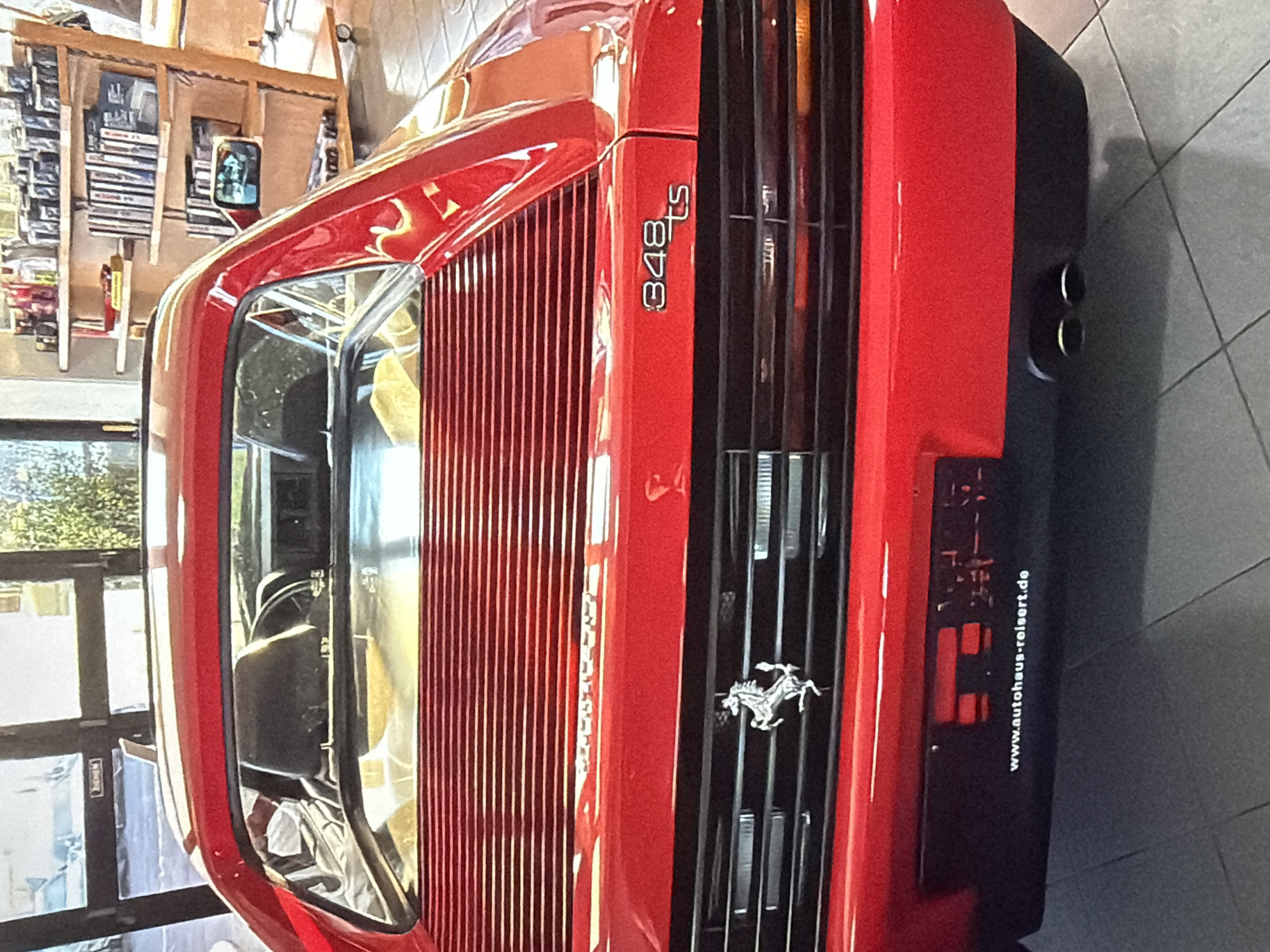 A rear view of a red Ferrari 348ts automobile is captured indoors. The car features the "348ts" badge and the Ferrari logo. In the background, an interior setting is visible, including shelves and what appears to be a window reflecting greenery outside. A sign with the text "www.autohaus-reisert.de" is attached to the car. The city of Burgas, Bulgaria is not discernible from the provided image.