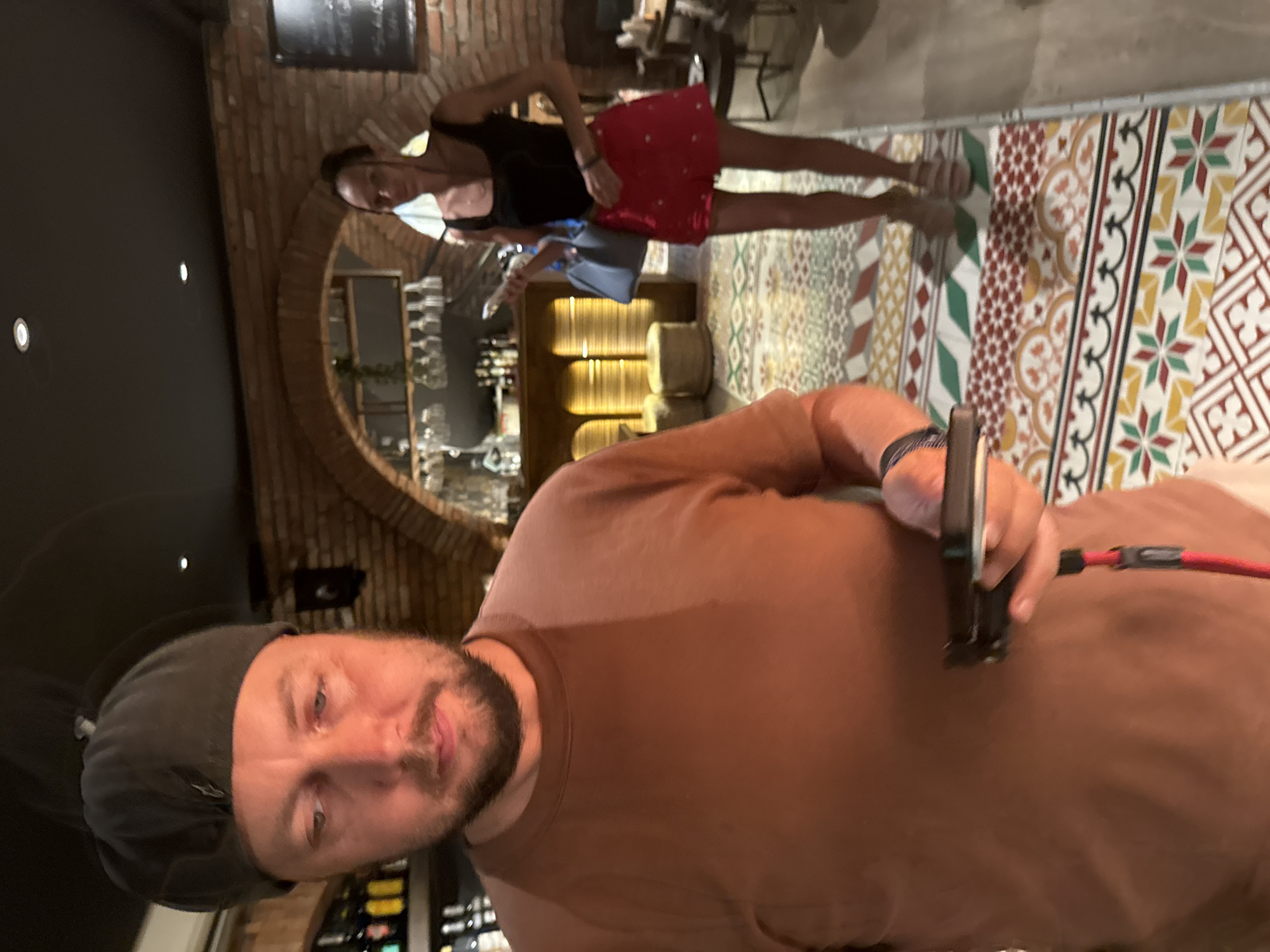 In an indoor establishment with brick walls and a tiled floor, a man in a brown t-shirt and a dark cap looks down at a phone in his hand. He has a beard and is in the foreground. In the background, a woman wearing a black tank top and a red skirt walks away from the camera, holding a light blue purse. The establishment appears to be a bar or restaurant, with shelves of bottles visible behind the man and wine glasses hanging from a rack behind the woman. The floor is decorated with a colorful geometric pattern. The lighting is dim, suggesting it is evening or nighttime. The location is identified as a bar in the city of Hanoi, Vietnam.