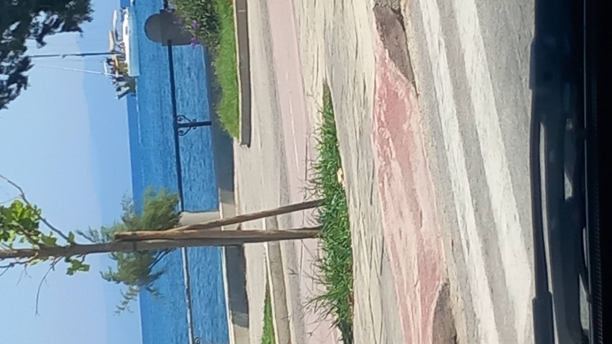 The image depicts a street scene with a pedestrian crossing in the foreground. Behind the crossing, there is a sidewalk with grass and a tree. In the background, the blue sea is visible with a sailboat on the water. The location is the city of Kos, Greece. The sky is clear and the weather appears to be sunny. The perspective is from a car, looking out through the windshield.