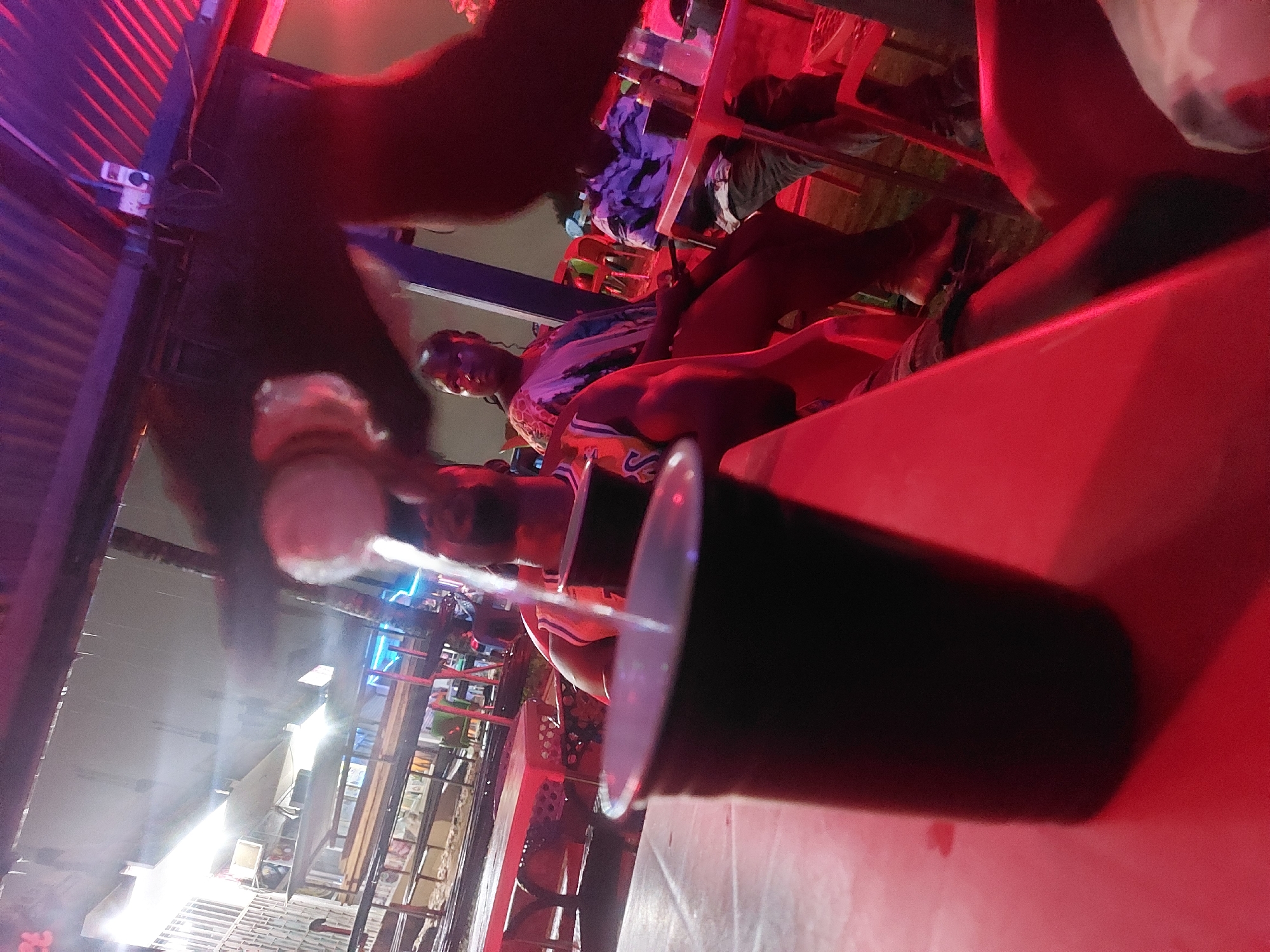 The image, taken in Dar es-Salaam, Tanzania, depicts a street-side scene at night, heavily illuminated by red lighting. A partially obscured foreground shows a plastic cup with a straw, suggesting someone is having a drink. Several people are visible, some seated and some possibly standing. Garments and other objects are scattered on nearby surfaces and furniture. A corrugated iron roof overhangs part of the scene. In the background, storefronts are visible, adding to the bustling atmosphere of the Tanzanian city.