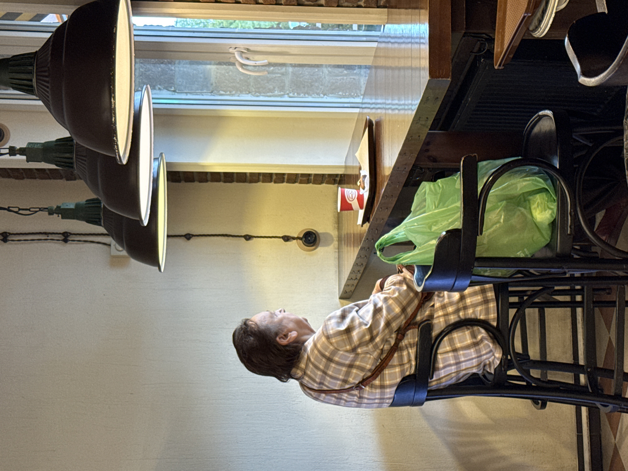 An elderly person with dark hair, wearing a plaid shirt, is seated at a bar in what appears to be a cafe or restaurant. They are looking to the right, out a window. On the counter in front of them is a tray with a red and white cup and a small pastry. A bright green plastic bag rests on the seat of the chair next to them. The cafe has industrial-style pendant lights hanging from the ceiling. The background shows a brick wall and a window with white handles. This scene is set in Vsevolozhsk, Russia.