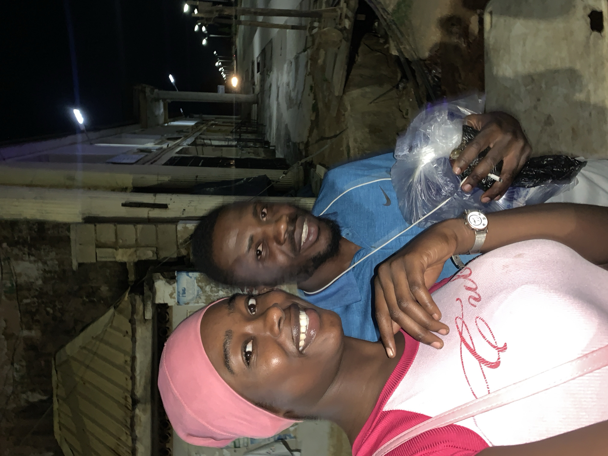 Here's a detailed description of the image:
**General Overview:** The image captures a close-up shot of two individuals, a woman and a man, in an outdoor setting at night. The background suggests a location in New Stadium, Nigeria, with evident infrastructure and urban features.
**People:**
* **Woman:** Positioned in the foreground, the woman wears a pink head covering and a pink top with red detailing. She's smiling, showing her teeth.
* **Man:** Situated behind the woman, the man wears a blue athletic shirt with a Nike logo. He is holding a clear plastic bag containing black items and is smoking a cigarette. He also is smiling.
**Objects:**
* **Plastic Bag:** Held by the man, the clear plastic bag contains dark, unidentifiable items.
* **Cigarette:** The man holds a lit cigarette.
* **Watch:** The man has a wristwatch on his left wrist.
* **Building Elements:** The backdrop features parts of buildings or structures that appear old and weathered. There are also lighting fixtures, possibly streetlights, and structural columns in the background.
**Environment:**
* **Nighttime:** The scene is set at night.
* **Urban Setting:** The presence of buildings, infrastructure, and streetlights suggests a populated, urban environment.
* **Location Clues:** The context provided indicates that the location is in New Stadium, Nigeria.
* Weathered: The buildings appear weathered, indicating the area may be subject to decay.
**Actions/Interactions:**
* The woman is looking directly at the camera and smiling.
* The man is holding a bag and smoking a cigarette, also smiling.
* The close proximity and smiles suggest a friendly or intimate interaction between the two individuals.