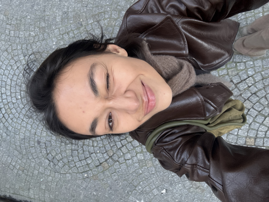 A young adult female is depicted in a high-angle, close-up shot, looking upwards at the camera. She has dark hair, dark eyes, and is winking her right eye while smiling with closed lips. Her skin tone is light to medium. She is wearing a dark brown, possibly faux leather, jacket with a wide collar and a visible zipper. Beneath the jacket, a textured, light brown or taupe scarf is wrapped around her neck. An olive green shoulder bag is visible over her left shoulder, with its strap and a portion of the bag showing. The background consists of a grey cobblestone pavement, arranged in a fanning or arc pattern, characteristic of many streets and squares in Paris, France. Scattered faint white specks, possibly small amounts of snow or ice, are visible on the cobblestones. A portion of a light brown or taupe shoe is visible at the bottom right, suggesting the subject is standing on the pavement.