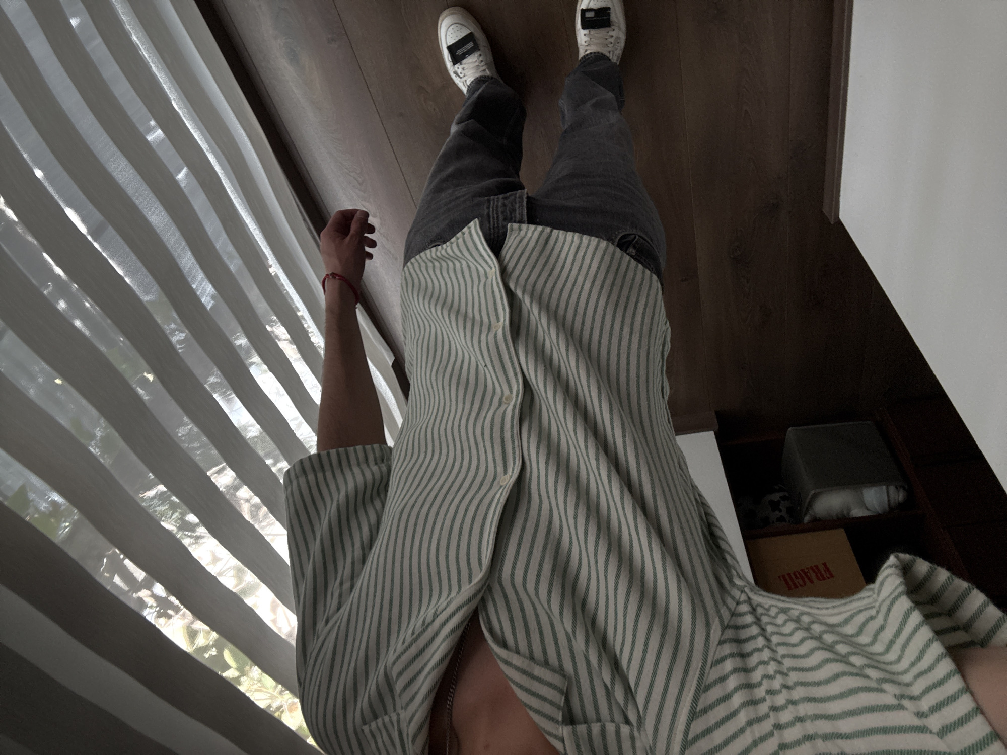 A first-person perspective shot from a standing position looking down at a person. The person is wearing white sneakers with a black tag on the tongue, gray jeans, and an unbuttoned, striped, light green and white shirt. The person's left arm is slightly raised and extended toward a window covered with white blinds. To the right of the person, a portion of a dark wooden floor is visible, along with a white wall and a cabinet containing a box with the word "FRAGILE" printed on it. The location of the image is within a building, but the exact city of San Bernardino Tlaxcalancingo, Mexico, is not verifiable from the image.