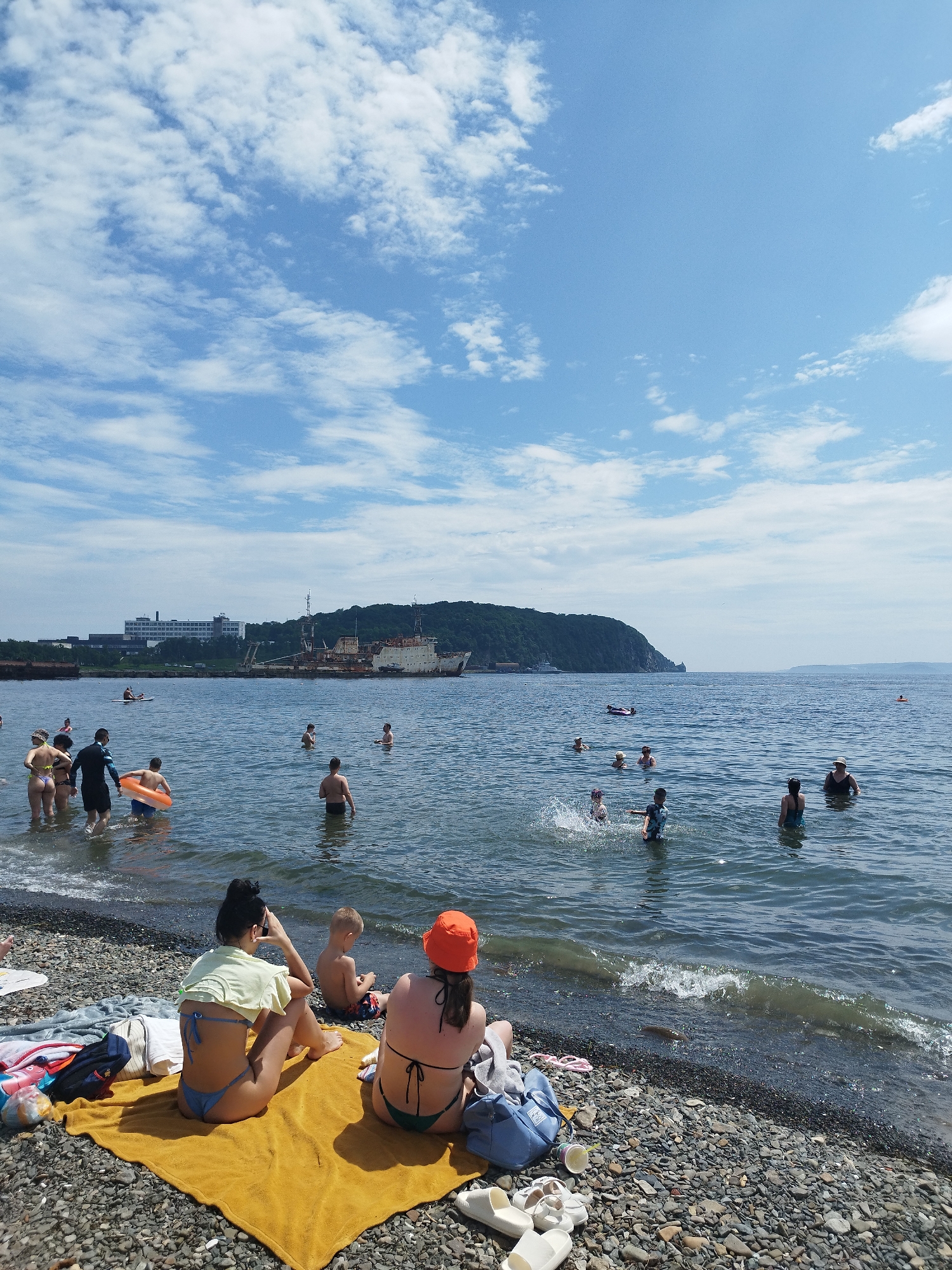 This media file depicts a lively scene at a pebbled beach on a sunny day. Several people are enjoying the water and the shore. In the foreground, two women are sitting on a yellow towel on the beach, with a young boy sitting between them. Further back, a group of people are wading in the shallow water, with some children playing and splashing. In the mid-ground, several adults are swimming and enjoying the ocean. A person is seen on a paddleboard in the distance.
The water is calm and clear, with small waves gently lapping the shore. The sky is a bright blue with scattered white clouds, suggesting fair weather. In the background, a large, rusted cargo ship is anchored in the bay, and a city or industrial area with buildings can be seen on the distant hills. The overall mood is relaxed and recreational, with people enjoying a summer day at the beach. The location is identified as Vladivostok, Russia. No visible text is present in the media file.