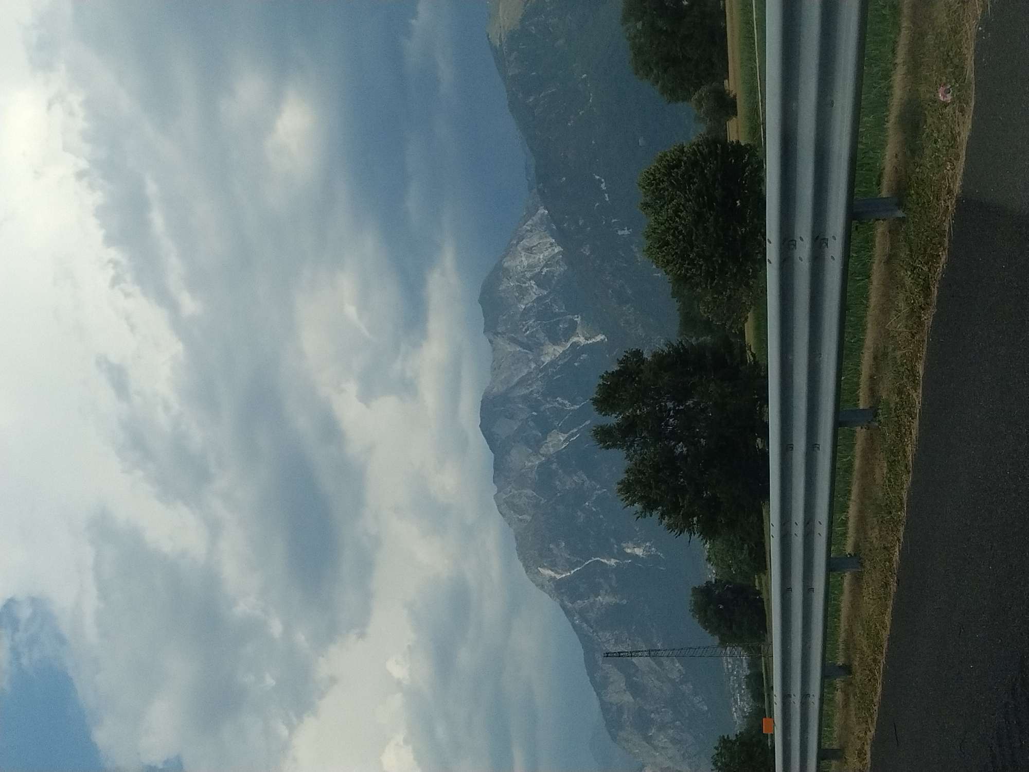 A wide shot captures a mountainous landscape under a cloudy sky, with a highway guardrail in the foreground. The mountains are rugged, with streaks of white suggesting rock or snow. In the middle ground, a dense line of trees separates the mountains from a field and the guardrail. A tall, metal structure, possibly a cell tower or power pylon, stands to the left of the mountains. The sky is filled with gray and white clouds, with a patch of blue visible in the upper left corner. The foreground shows a dark asphalt road, a dry grassy verge, and the silver guardrail. The scene is likely located in the outskirts of Buja, Italy, a town known for its mountainous terrain. There are no people or discernible actions visible in the image.