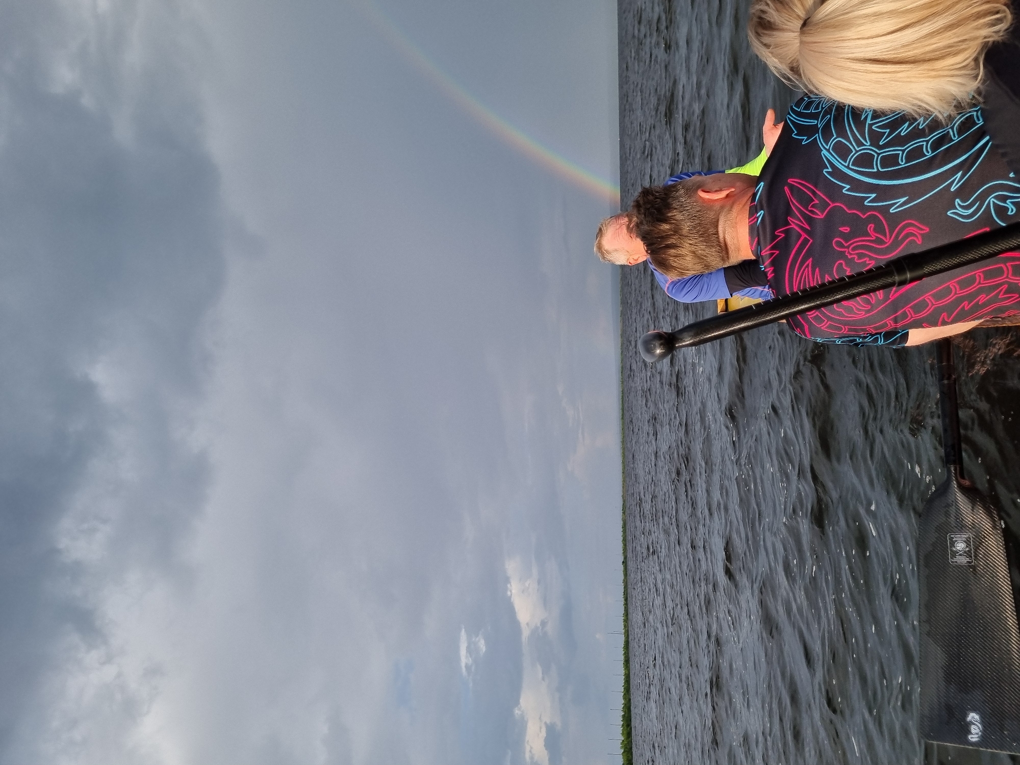 The image shows a group of people on a boat on a body of water under a cloudy sky with a rainbow. Two individuals are visible from behind, with one wearing a black t-shirt with a neon dragon design in pink and blue. Another person, an older male with gray hair, is partially visible looking forward, wearing a blue shirt. A paddle is positioned vertically on the right side of the frame. The background shows a distant shoreline with vegetation and structures, possibly indicative of the Kama River in Chaikovsky, Russia. The water is choppy, and the overall atmosphere is overcast with the presence of the rainbow suggesting a recent or ongoing rain shower.