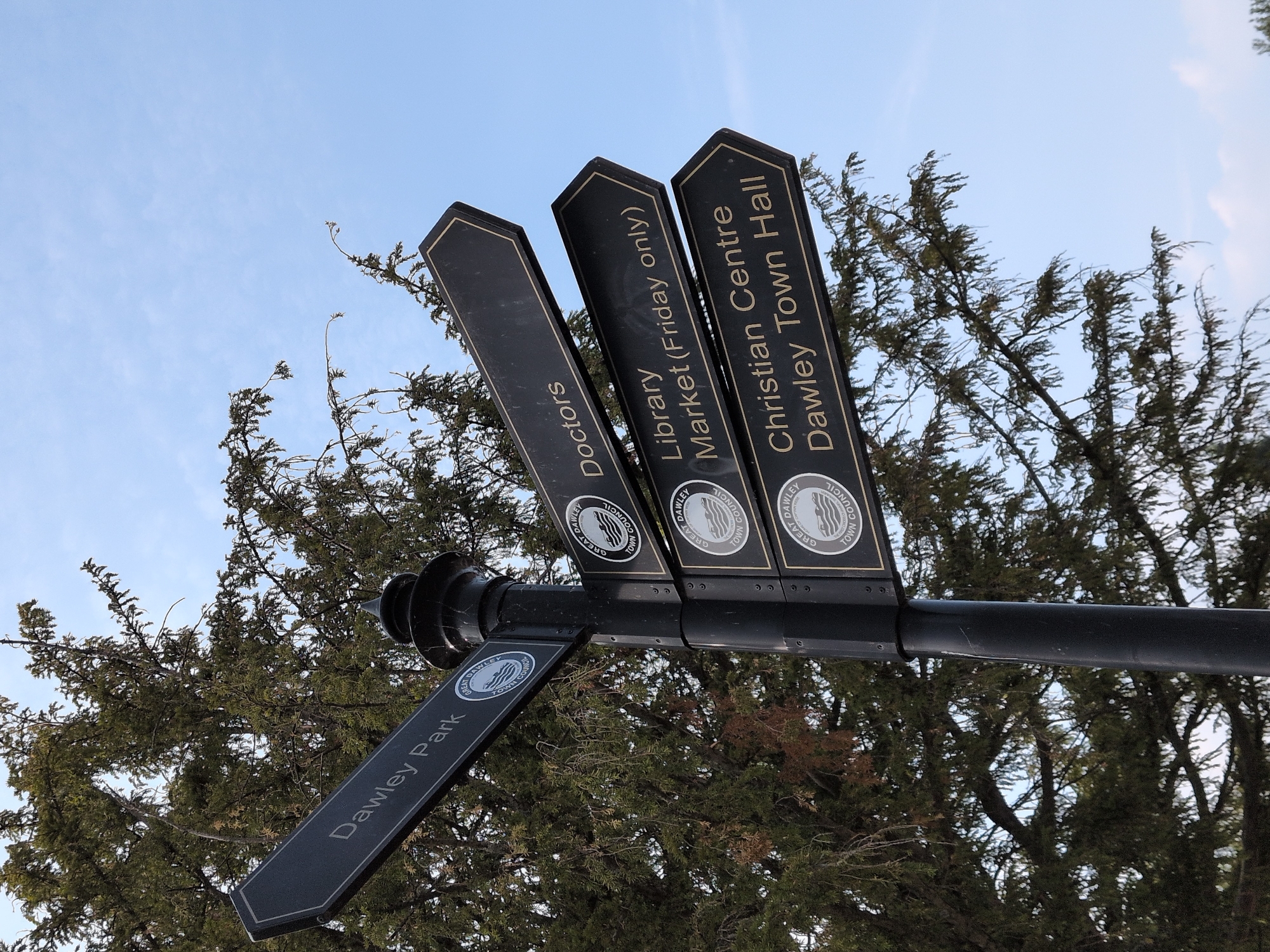 A black metal signpost stands against a partly cloudy sky, partially obscured by the branches of a large evergreen tree. The signpost has multiple directional arms pointing left and right, each bearing white text and a circular emblem with "GREAT DAWLEY TOWN COUNCIL" around its circumference.
The uppermost arm on the left points left and reads "Dawley Park". Below this, on the right side of the post, a series of arms point to the right. The topmost of these reads "Doctors". The next arm down says "Library Market (Friday only)". The lowest visible arm reads "Christian Centre Dawley Town Hall". The emblem on all the arms is consistent.
The signpost is located in Dawley, United Kingdom, a historic market town in Telford and Wrekin, Shropshire. The sign is a public amenity providing directions to local facilities and attractions within the town. The overall impression is of a clear, sunny day in a well-maintained public space.