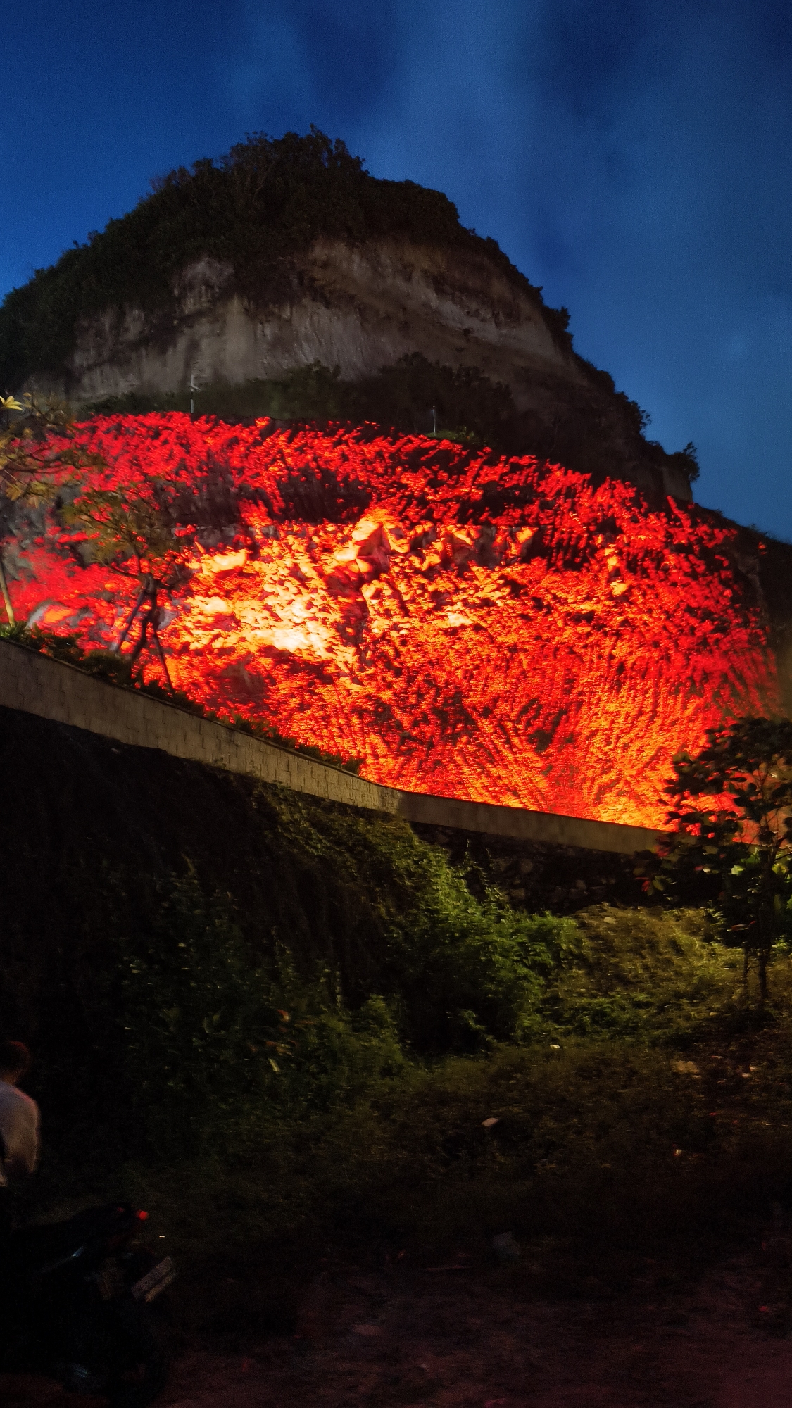 At night in Ungasan, Indonesia, a large cliff face is intensely illuminated with red light. The cliff dominates the scene, with a vegetated top and a sheer, rocky face that is brightly lit, creating a dramatic visual effect. A low retaining wall runs horizontally across the cliff's base, separating it from a slope covered in dense green foliage. In the lower portion of the image, the vegetation and surroundings are dark, with only hints of details. A person is partially visible on the lower-left side, with what appears to be a motorcycle parked nearby. The sky above the cliff is a deep blue with faint cloud formations.
