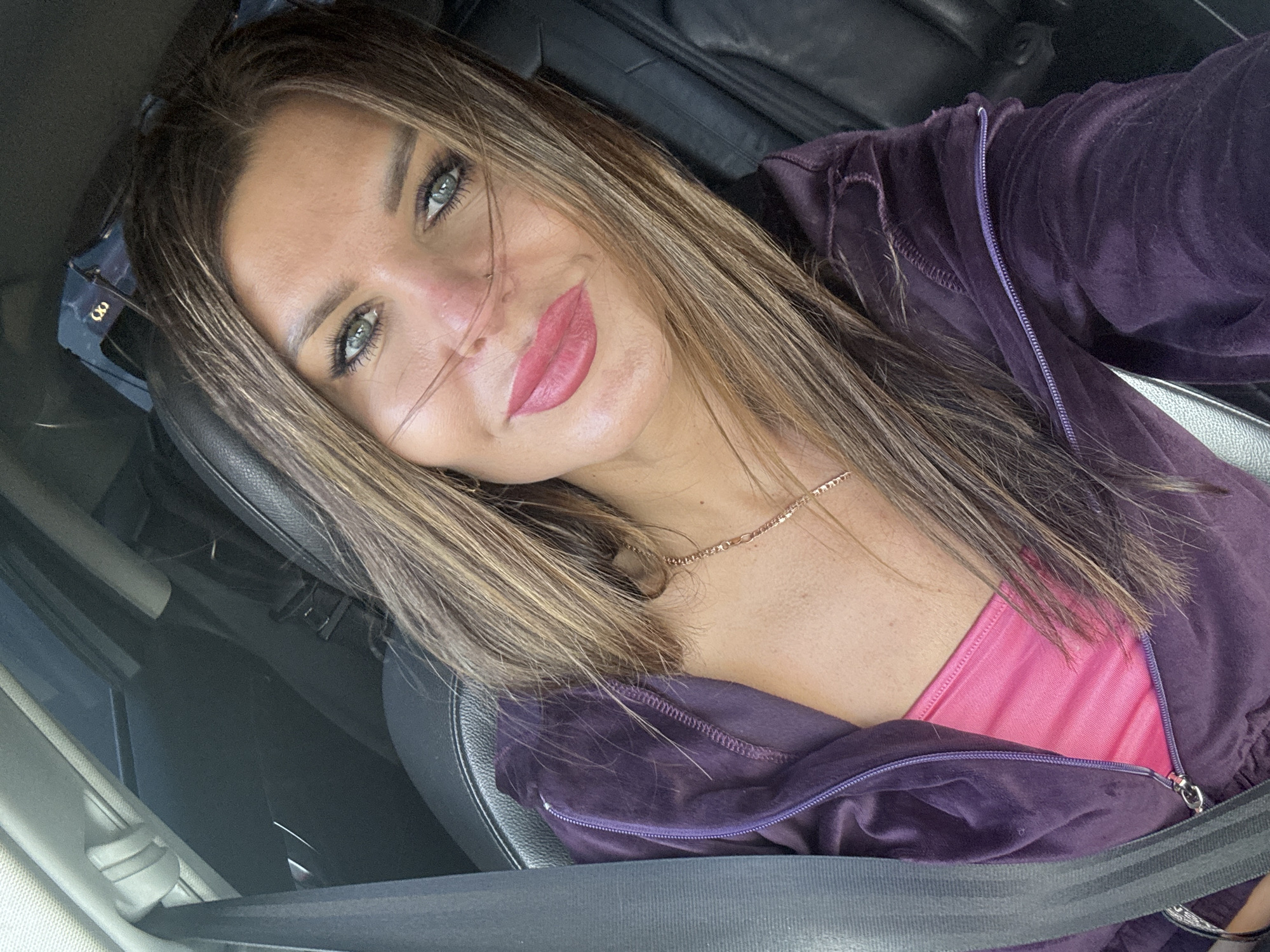 A medium shot from a first-person perspective depicts a woman seated in the driver's seat of a passenger vehicle, looking directly at the camera with a slight smile. She has long, straight, light brown hair with lighter highlights, prominent green eyes accented with dark eyeliner and mascara, and defined eyebrows. Her lips are full and colored with matte pink lipstick. She is wearing a dark purple velour or velvet hooded jacket, partially unzipped to reveal a bright pink top underneath. A thin, gold-colored chain necklace with small decorative elements is visible on her upper chest. A grey-black seatbelt diagonally crosses her chest and is fastened. The car's interior features black leather seats and dark trim. In the upper right background, a pair of dark sunglasses with a gold "CC" or infinity-loop style logo is visible, likely resting on the headliner or rear parcel shelf. Further back, a dark object, possibly a bag, is discernible on the rear seat. The lighting is bright and diffuse, indicating daytime. The generic appearance of the car's interior, devoid of specific landmarks or identifying markers, does not provide any evidence to determine a precise geographical location, such as Chaikovsky, Russia, or any other specific city or country.