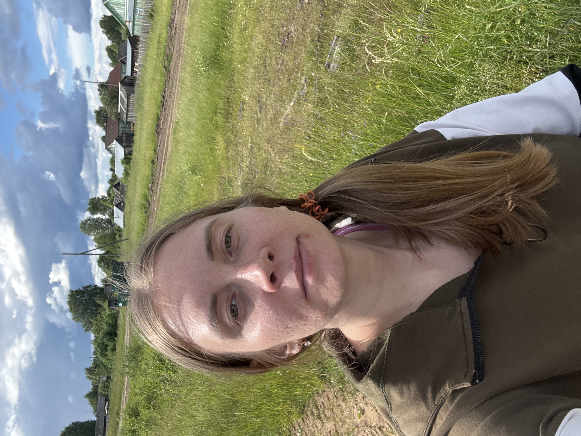 This is a vertical, close-up selfie of a young woman with blonde hair, taken outdoors on a sunny day. She is wearing a dark olive-green jacket with white and black accents on the sleeves. Her hair is pulled back with an orange hair tie. She has a light complexion and is looking directly at the camera with a neutral expression.
The background suggests a rural or village setting in Kurgan, Russia. A dirt path runs through tall, green grass, with scattered small wildflowers. Beyond the grass, a line of traditional wooden houses with peaked roofs and wooden fences is visible. Trees are scattered throughout the landscape. The sky is a bright blue with fluffy white clouds. The lighting suggests it is daytime, likely late morning or afternoon, with bright sunlight illuminating the scene. No text is visible in the image.