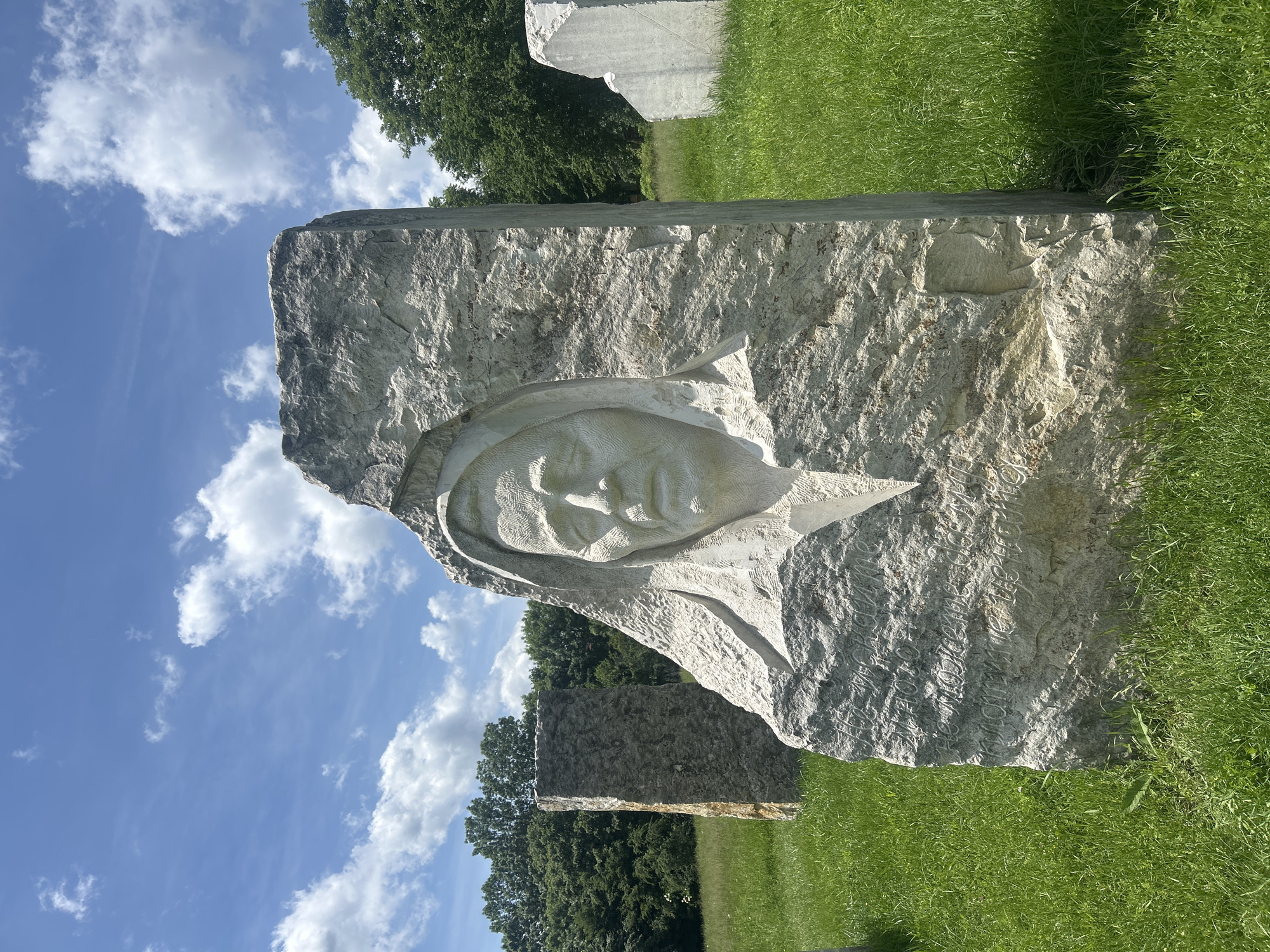The image features a large, light-colored stone sculpture prominently displayed outdoors in what appears to be an art park or memorial site, located in Gardevtsi, Bulgaria. The main subject is a substantial, rough-hewn stone block, carved in bas-relief with the serene face of a woman, possibly wearing a head covering or veil. Her expression is calm and contemplative. The texture of the natural stone is evident, with the carved features showing a smoother finish.
Below the carved face, on the lower part of the stone block, an inscription is visible. The engraved text is in Cyrillic script and can be partially deciphered as "СПОМЕНЪТ ЗА НЕЯ БЕЗСМЪРТЕН...", which translates to "The memory of her is immortal...". This suggests the sculpture may be a memorial piece.
The setting is a vibrant green grassy field, with several other large stone blocks or unfinished sculptures scattered in the mid-ground, indicating a broader sculptural installation. Beyond the field, a line of dense green trees forms a natural backdrop. The sky above is clear and blue, dotted with fluffy white clouds, suggesting a sunny day, likely midday or early afternoon, given the bright, direct lighting. No specific activity is depicted, but the scene evokes a sense of tranquility and reflection. The original image is captured with a vertical orientation, rotated 90 degrees counter-clockwise.