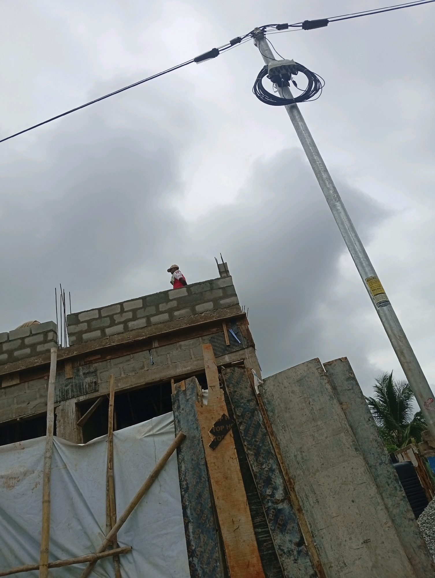 In Akowonjo, Nigeria, a construction site is visible under a cloudy sky. A tall, gray metal utility pole with coiled black cables and a junction box stands prominently on the right side of the image. To the left, a building under construction features concrete block walls with visible rebar. A person wearing a red shirt and a cap is standing on top of the partially built wall, appearing to be working. Wooden scaffolding and white tarps cover parts of the structure. Plywood boards and construction debris are scattered around the site. A palm tree is visible in the background.