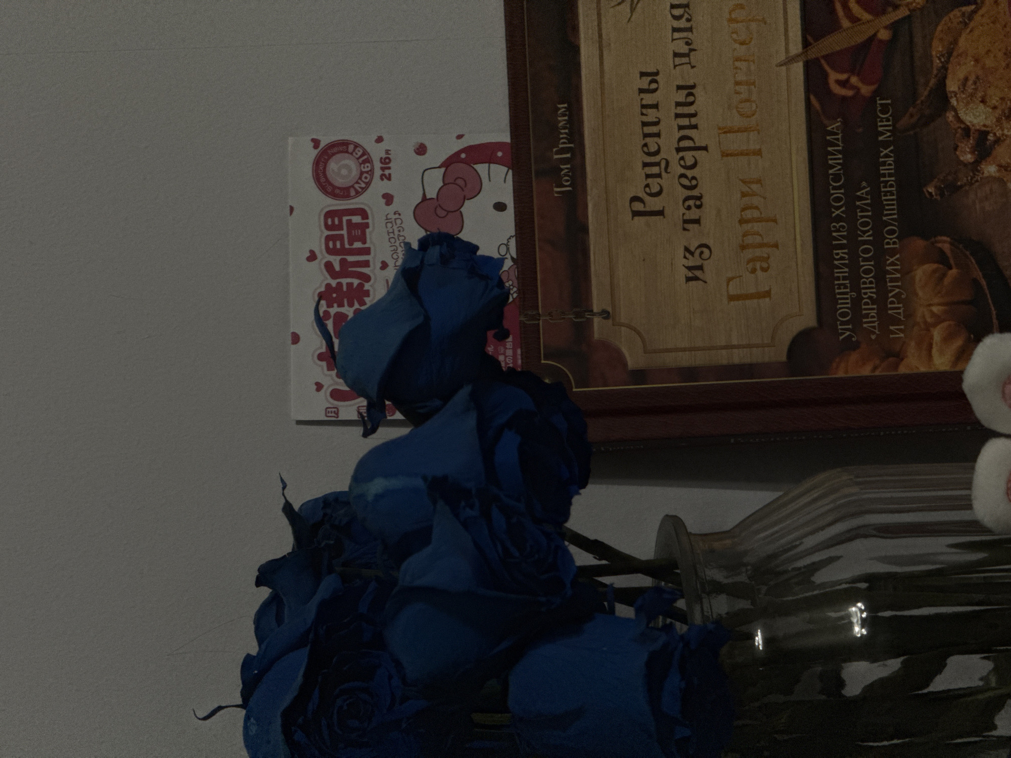A close-up, dimly lit indoor shot features a bouquet of vibrant blue roses in a fluted glass vase. Behind the roses, a partially visible, pink and white box with Japanese text and a Hello Kitty illustration is present. To the right, a book with a wooden-textured cover prominently displays Russian text that translates to "Recipes from taverns for Harry Potter". The lighting is low, casting shadows and highlighting the textures of the flowers, vase, and book. No specific location within Sinyavino, Russia, is identifiable from the visual cues. There are no people or discernible actions taking place.