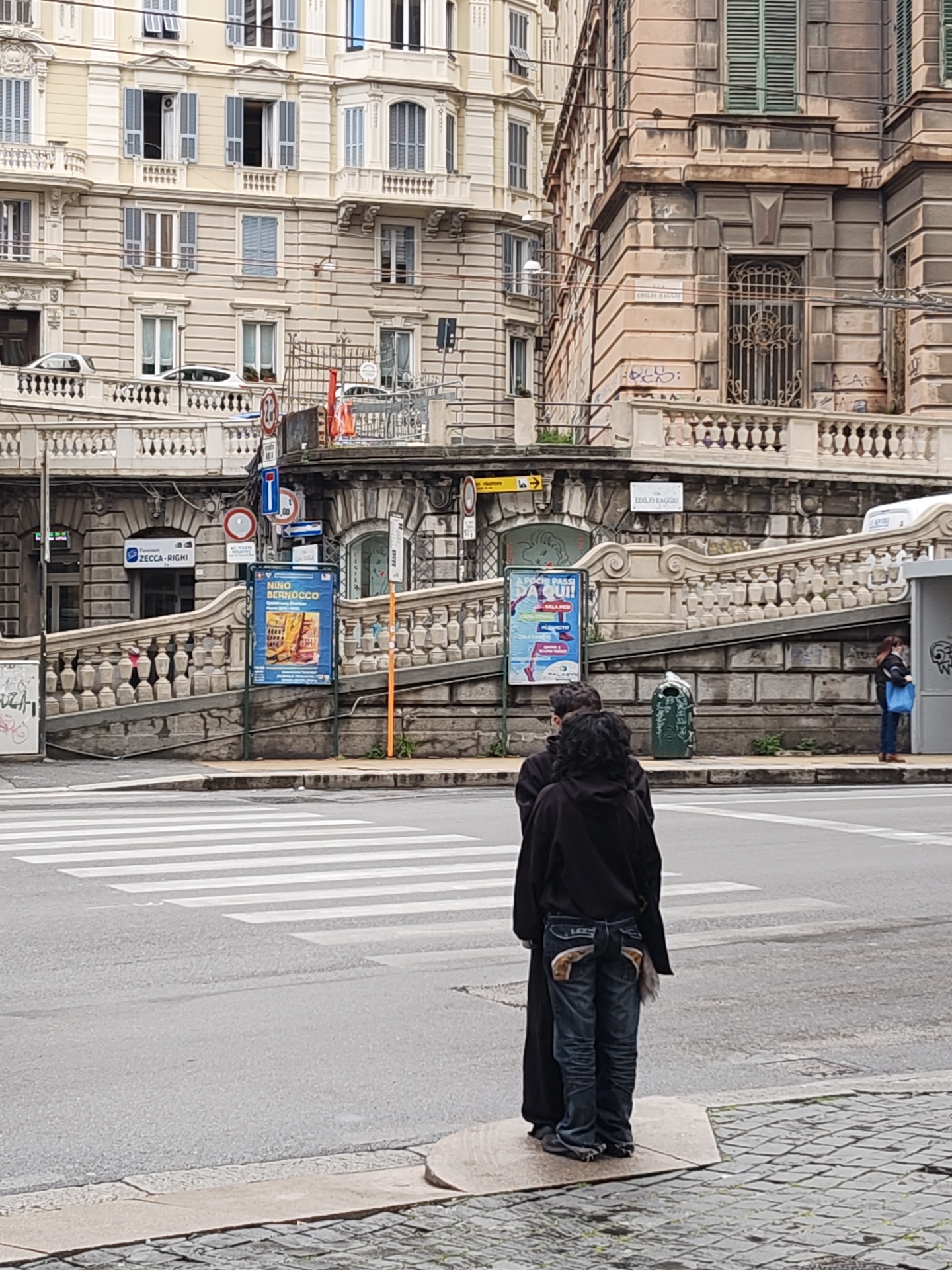 The image depicts a street scene in Genoa, Italy, specifically near the Piazza Dante and the steps of the "Scalinata Zecca-Righi" (Zecca-Righi staircase), indicated by the "ZECCA-RIGHI" sign. Two people are standing on a section of the sidewalk beside a crosswalk, with their backs to the viewer. One is wearing a black hooded coat, and the other has jeans with detailed back pockets and a tail on the lower back. In the background, are buildings with architectural details typical of Genoa, including ornate balconies and window shutters. There are traffic signs and posters on display. A bus stop is partially visible on the right side of the image, where a person wearing a mask is waiting. A green trash container is on the sidewalk next to the staircase. The street and sidewalk are wet, suggesting recent rain. The image is taken at eye level.
