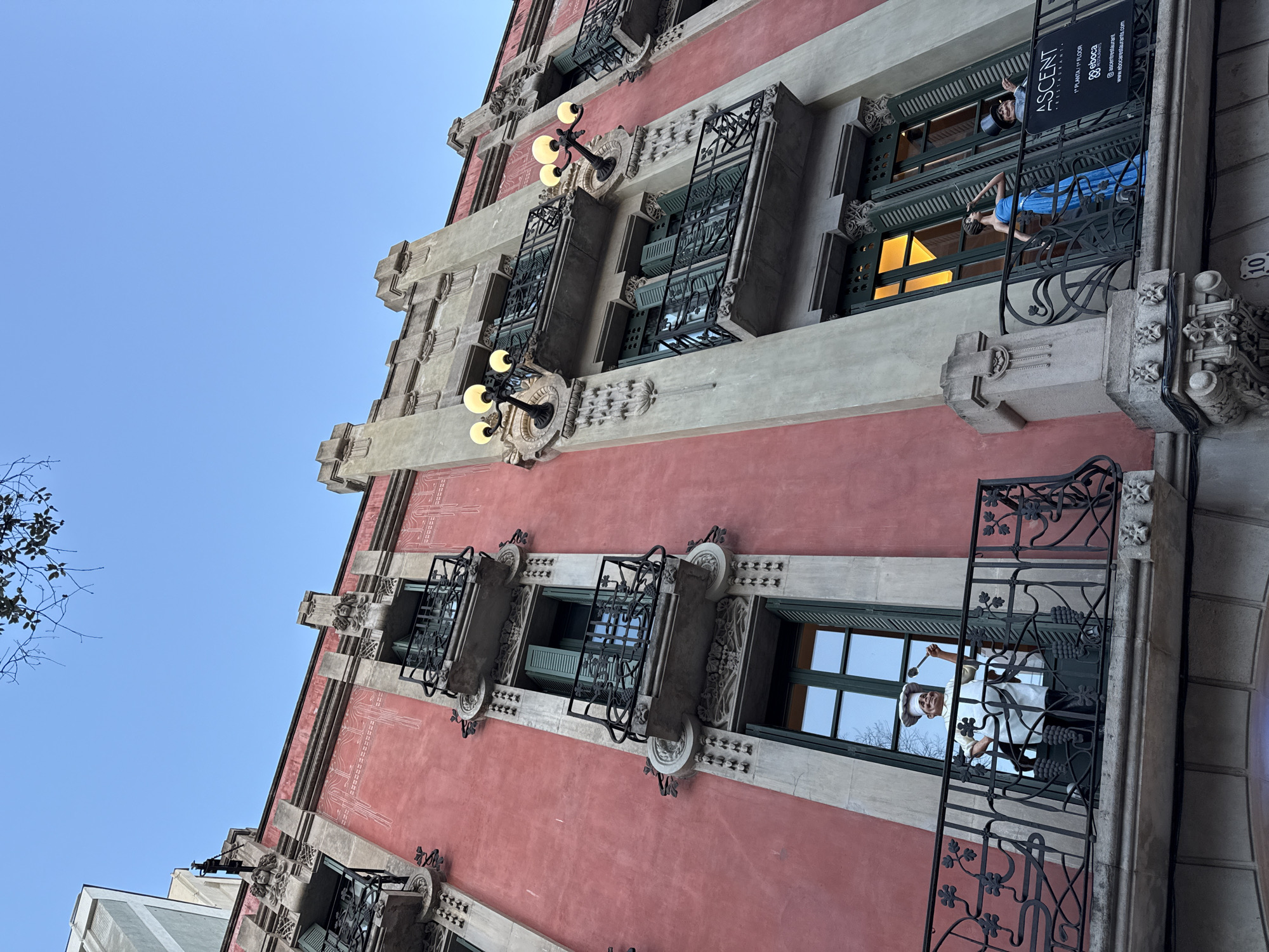 A low-angle shot of a building facade in Barcelona, Spain. The building is primarily a pale pink color with architectural details in grey. There are several windows with ironwork balconies. On one of the balconies, there is a figure dressed in blue, likely a mannequin or sculpture. Above this balcony, a sign hangs advertising "ASCENT Restaurant, 1st Floor." Another balcony features a figure dressed as a chef. The overall scene captures the architectural style and character of Barcelona. The sky is a clear light blue.