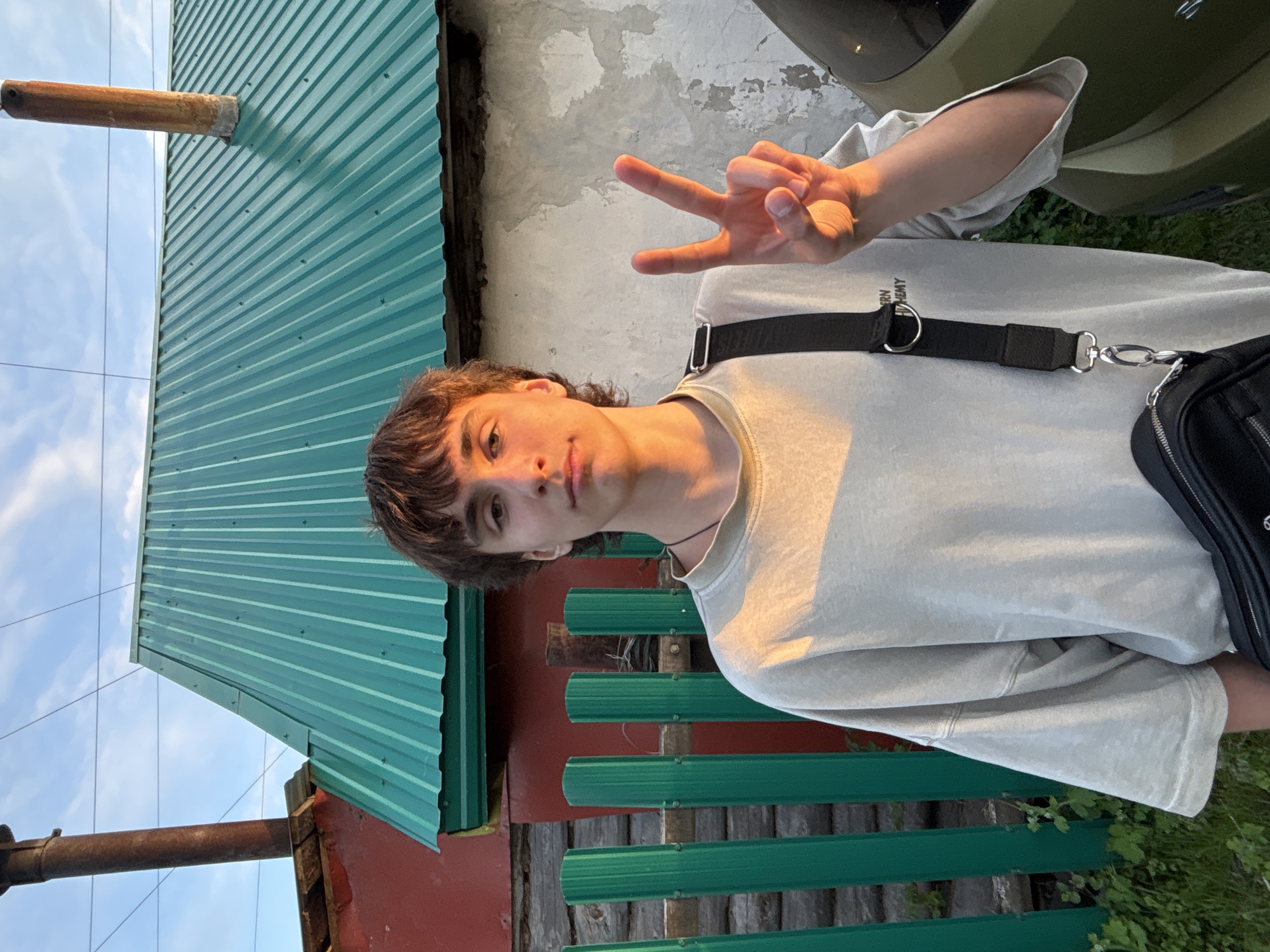 A young man stands outdoors making a peace sign with his right hand. He is wearing a light grey t-shirt and a black shoulder bag. The background includes a building with green corrugated metal siding, a fence, a vehicle, and the sky. The location is likely Chaikovsky, Russia, given the prompt.