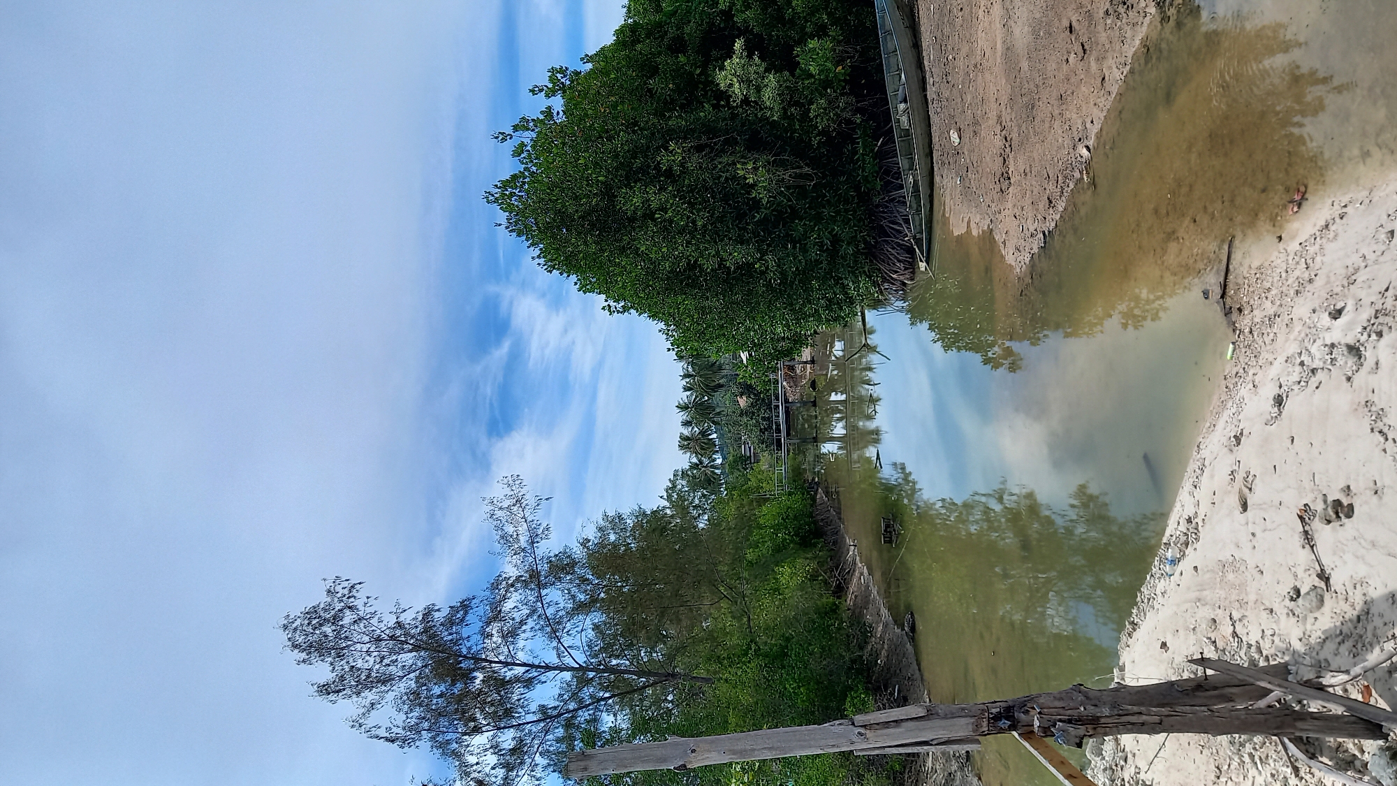 The image depicts a serene tropical waterway in Tambon Koh Phangan, Thailand. The water is calm and reflects the sky and surrounding lush vegetation, creating a mirror-like effect. On the left bank, a weathered wooden post stands prominently, with a dense growth of trees and shrubs behind it. The right bank is characterized by sandy soil and features a narrow, elongated boat resting on the shore. Further down the waterway, a small bridge can be seen, with more trees and palm trees visible in the distance, suggesting a coastal or riverine village setting. The sky is partly cloudy, with bright blue patches interspersed with white, wispy clouds. The overall scene conveys a sense of tranquility and natural beauty characteristic of a tropical environment.