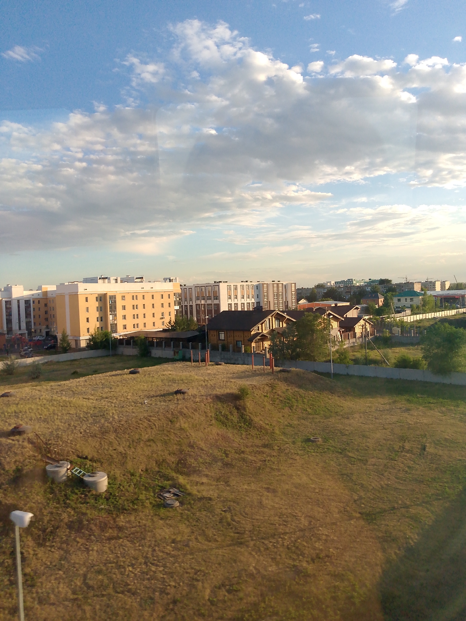 This image depicts an urban landscape under a partly cloudy sky, likely in Astana, Kazakhstan. The scene captures a blend of residential buildings and undeveloped land. In the foreground, a grassy, undulating hill is dotted with some circular concrete structures, possibly remnants of construction or water tanks, and a fallen ladder. A street light stands on the left side of the frame.
Further back, a row of modern apartment buildings dominates the middle ground. These structures are characterized by their multi-story design, with visible windows and balconies, and present a mix of beige, white, and darker facade colors. Adjacent to these apartments, a cluster of wooden houses with prominent gabled roofs suggests a more traditional or suburban residential area. Fences and greenery are visible around these homes.
In the background, more buildings of various sizes and architectural styles can be seen, extending towards the horizon. Cranes are visible in the distance, indicating ongoing construction or development in the area. The sky is a mix of blue and white clouds, with the sun casting a warm light, suggesting it is either early morning or late afternoon. There are no people clearly visible in the image, and the overall impression is one of a developing urban area transitioning between open space and new construction. No visible text is present.
