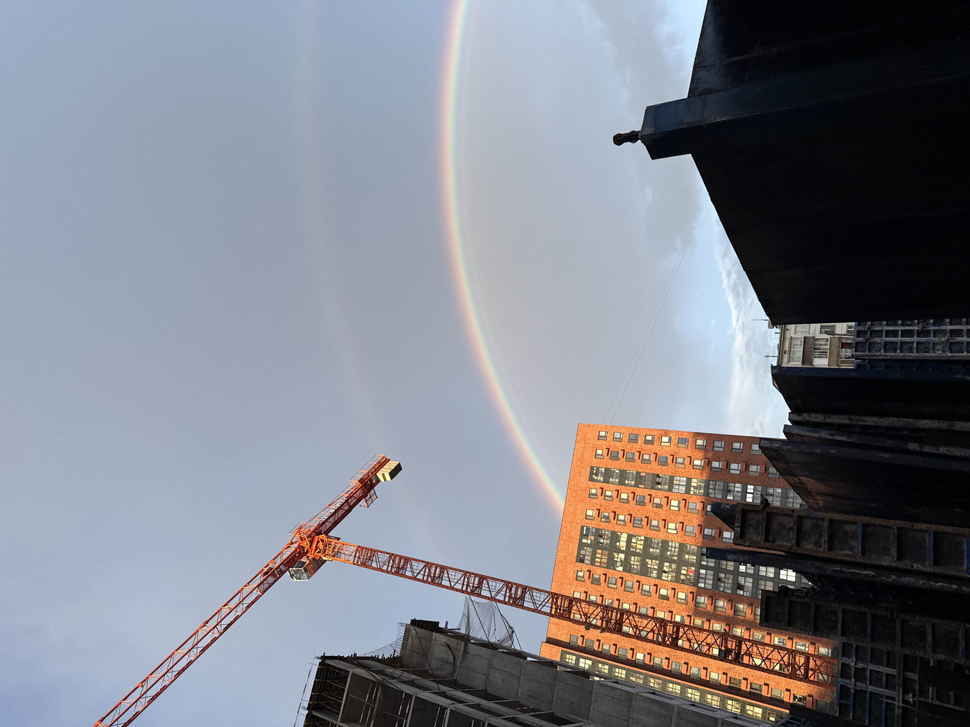 The image depicts a construction site in Moscow, Russia, under a sky with a visible double rainbow. In the foreground, the dark, angular structures of unfinished buildings or construction materials dominate the lower portion of the frame. To the left, a tall, orange construction crane is prominently positioned, extending its arm upwards and to the right. Adjacent to the crane, a partially constructed building, appearing to be made of concrete and rebar, rises on the left side. In the middle ground, a completed, multi-story residential building with a red brick facade and numerous windows stands illuminated by sunlight. The sky above is partly cloudy, with the faint arc of a secondary rainbow visible above the primary one. The scene captures a moment of contrast between ongoing urban development and a natural atmospheric phenomenon.