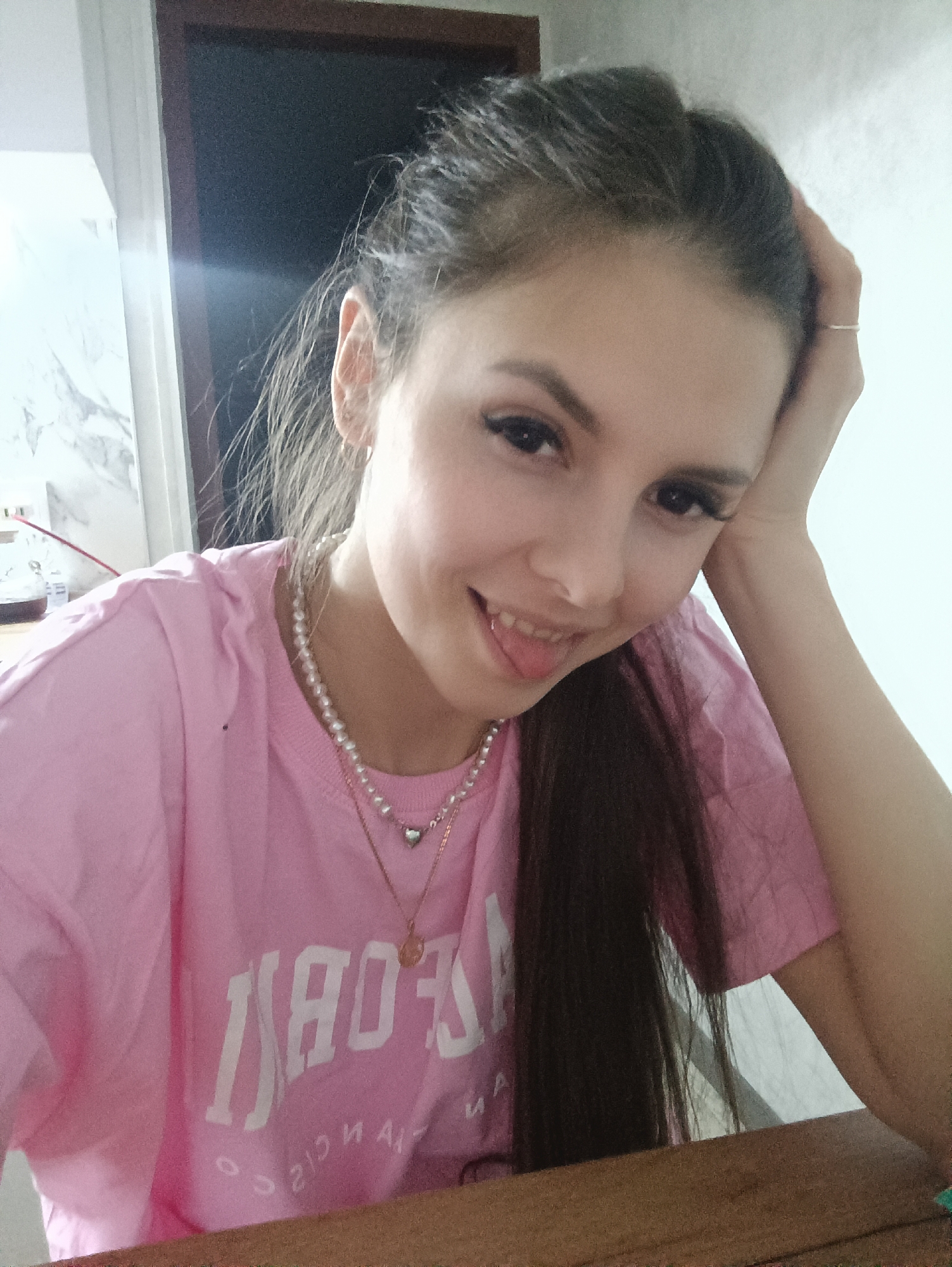 A woman with brown hair pulled back and wearing a pink T-shirt with white lettering is posing for a selfie. She is smiling and sticking out her tongue slightly. She is wearing a pearl necklace with a heart charm and a gold necklace with a pendant. Her left hand is resting on her head. She is sitting at a wooden table. In the background, there is a doorframe and a light-colored wall. The location is Unknown, Unknown.