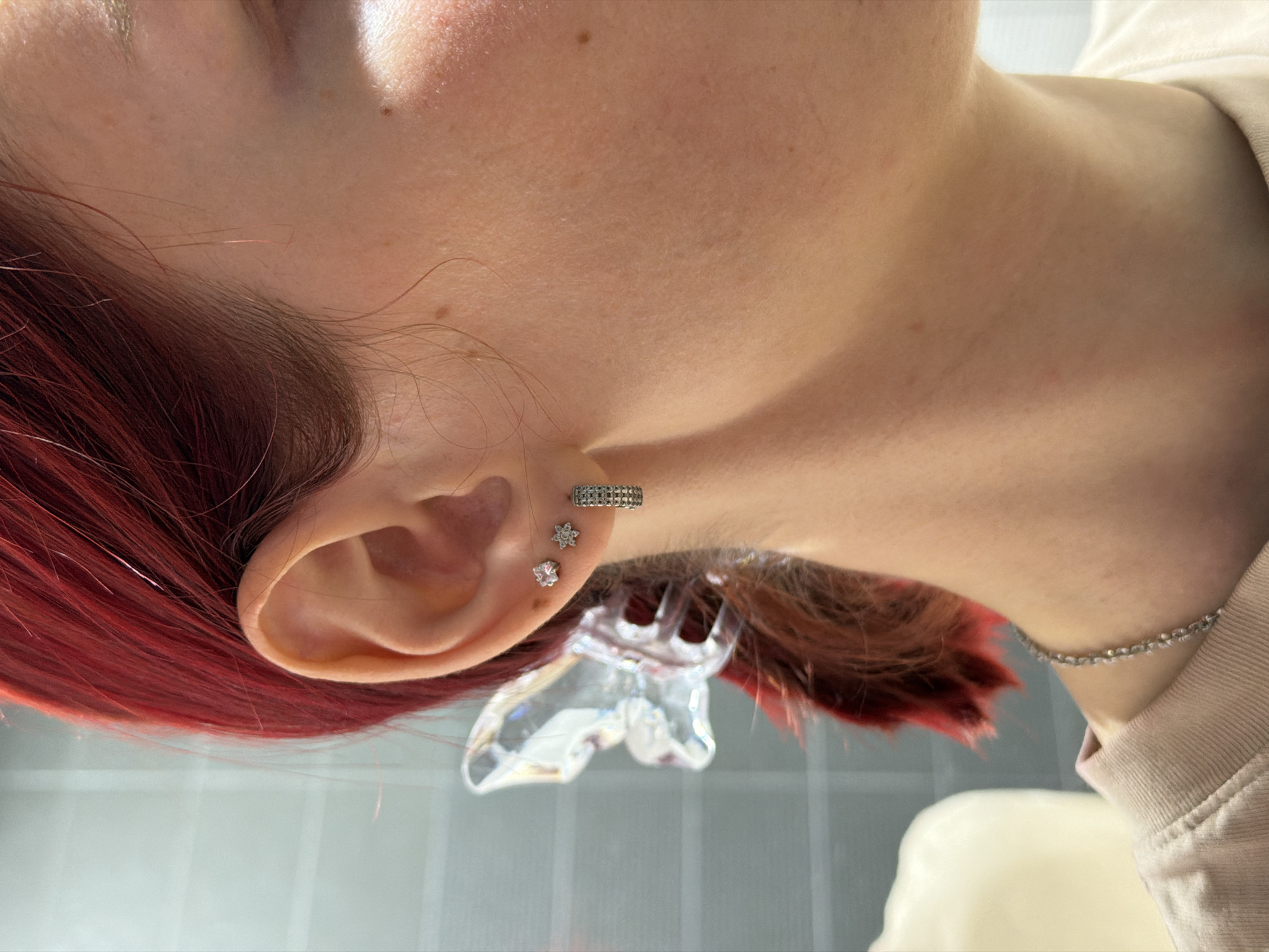 A close-up, tilted-perspective image shows the side of a person's head, likely a female, with red-dyed hair. The focus is on the left ear, which is adorned with three pieces of jewelry: a hoop earring at the top of the ear, and two gemstone studs, one larger than the other. A small, clear decorative butterfly or wing-shaped object is positioned near the ear, partly obscured by hair. A silver chain necklace is visible on the neck. The background is a light-colored surface, possibly tile, with a grid pattern. The location is unknown.